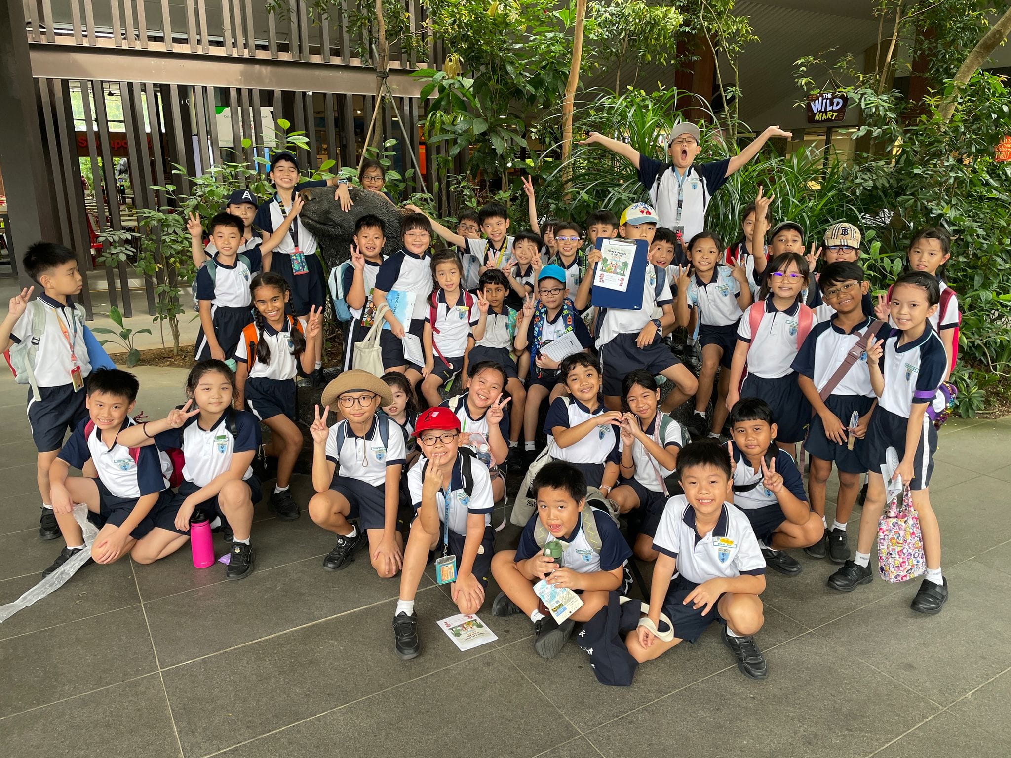 A large group of young students in white and navy uniforms pose cheerfully outdoors near green plants.