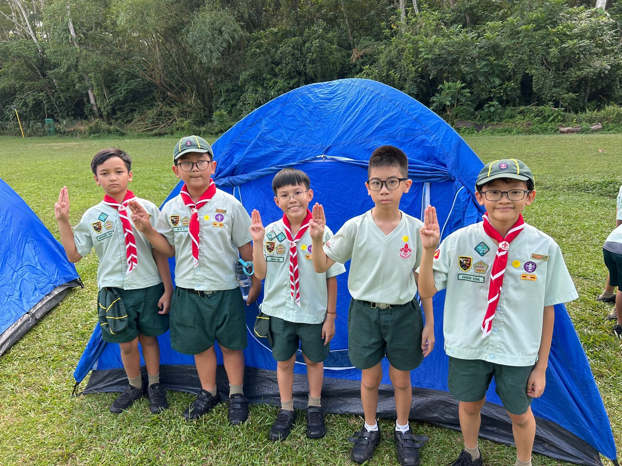 Five boys in scout uniforms with red neckerchiefs give the scout salute in front of a blue dome tent on grass.