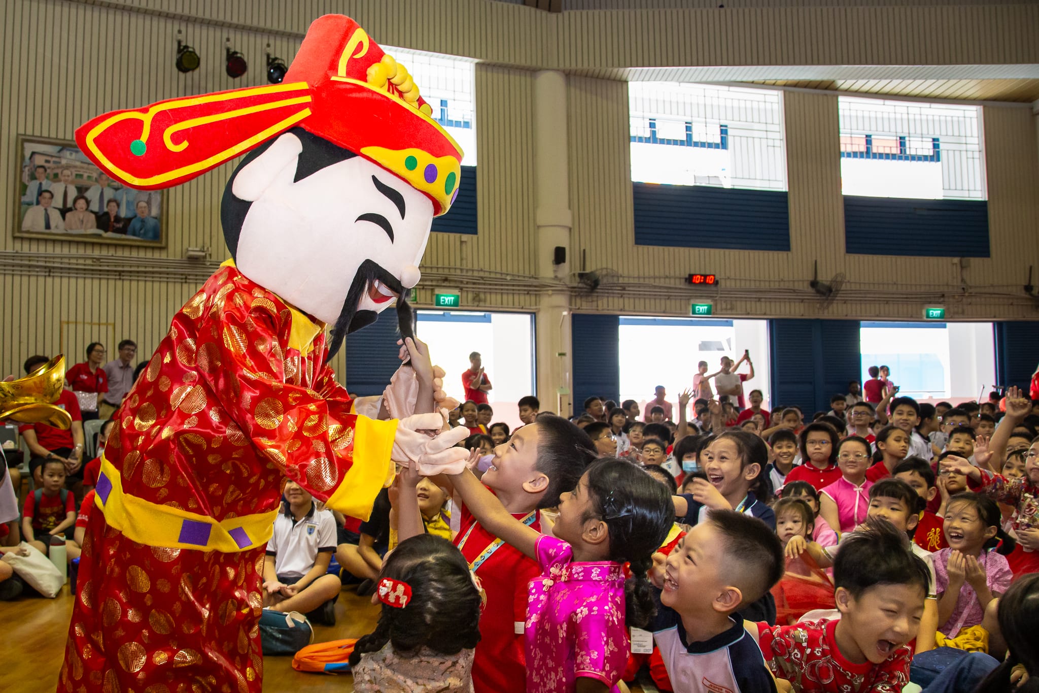 A Chinese opera mascot in red costume interacts with excited children in a school hall.