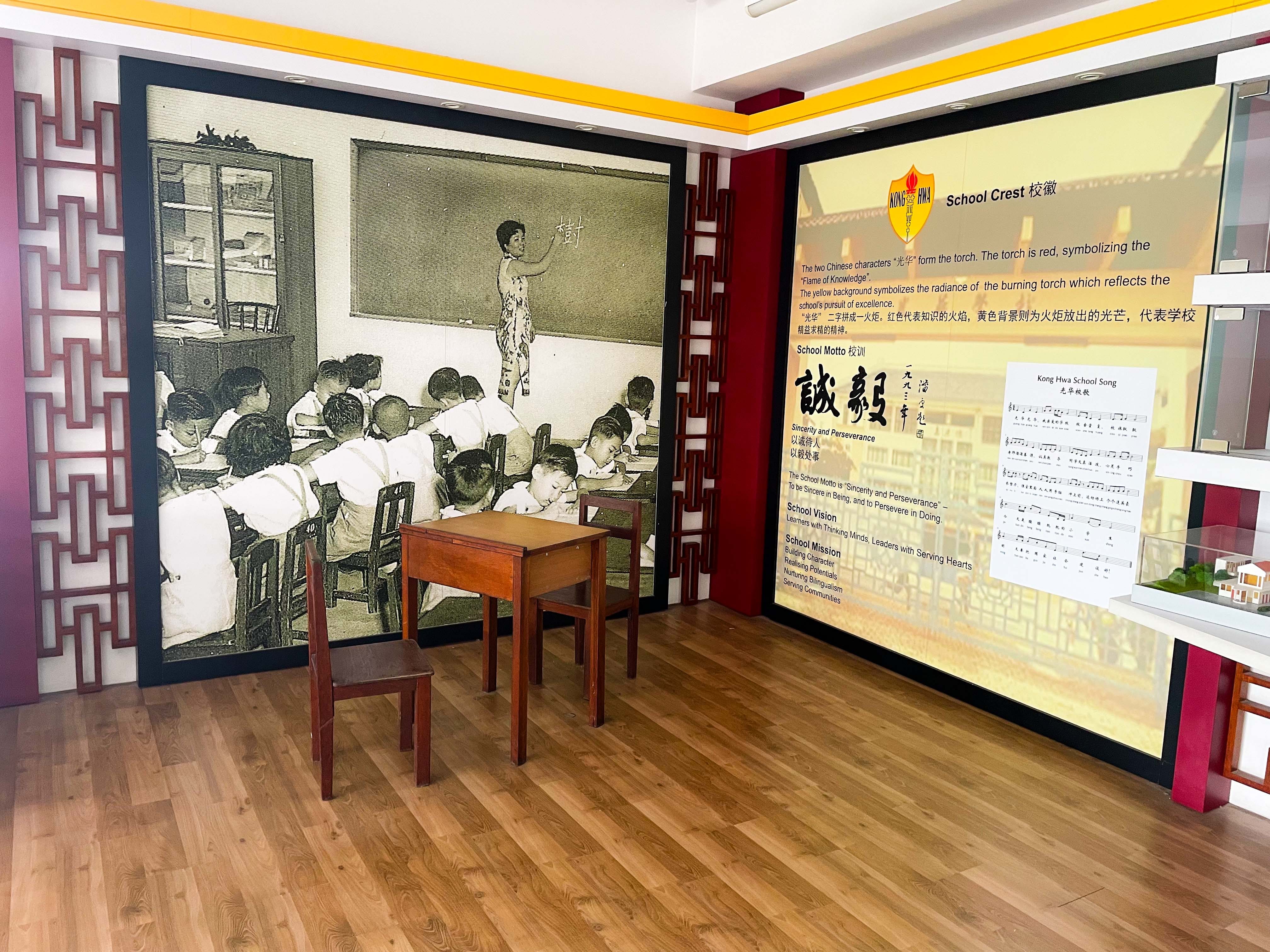 A museum display with a vintage classroom photo mural, a Kong Hwa School crest panel, and a small wooden desk with chairs.