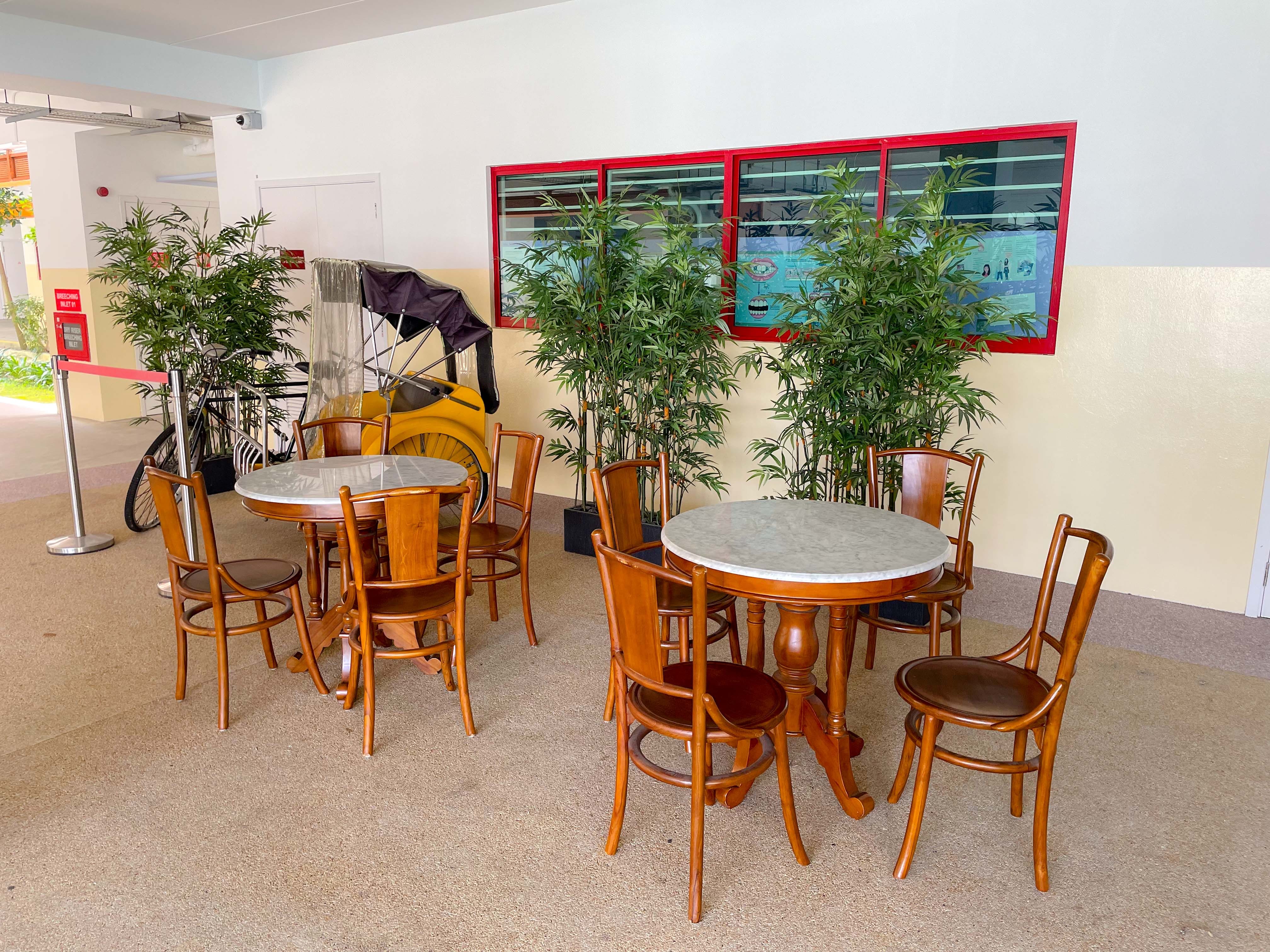 Two round marble-top wooden tables with chairs, green potted plants, and a vintage trishaw against a cream wall.