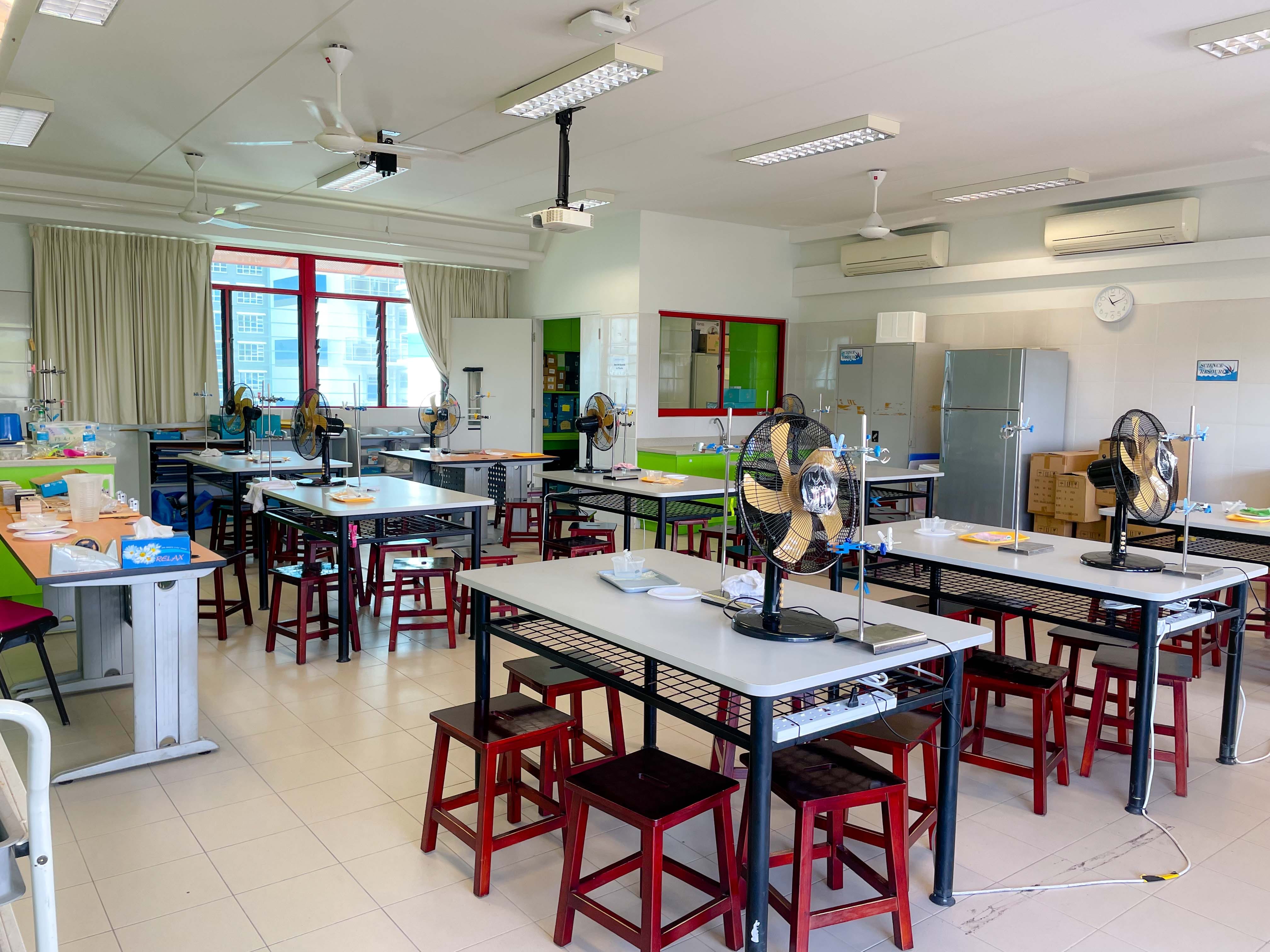 Empty school science lab with gray workbenches, red stools, desk fans, retort stands, ceiling projector, and air conditioners.