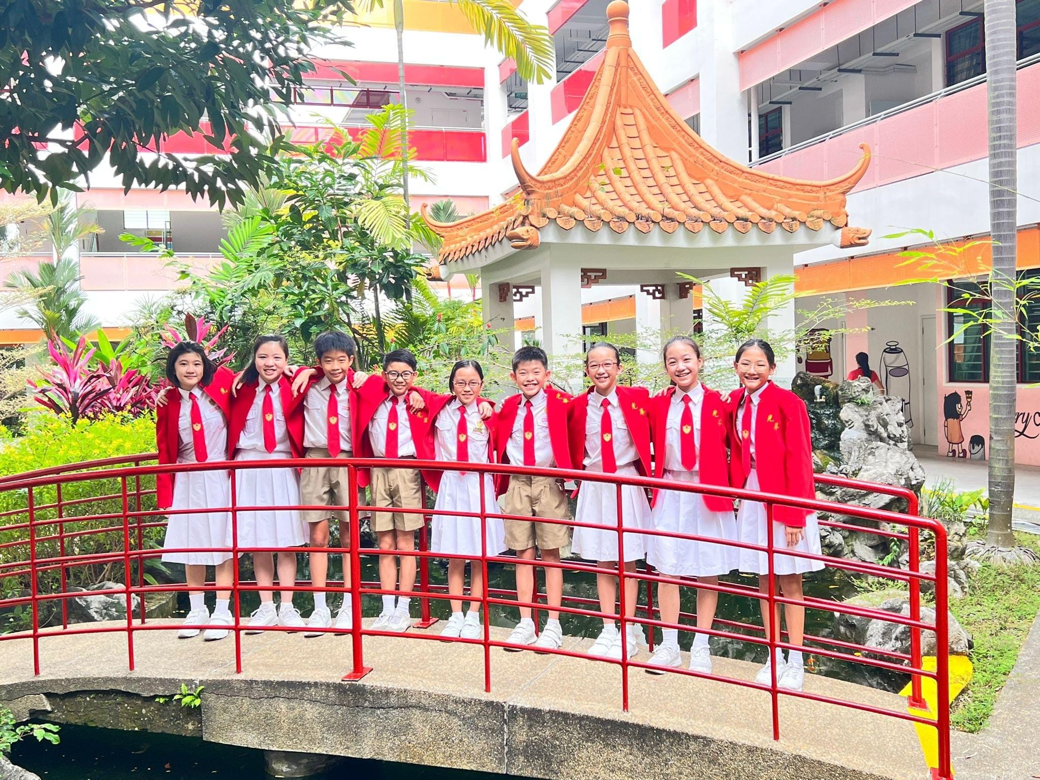 Eight children in red blazers and uniforms stand smiling on a red arched bridge near a Chinese pagoda.