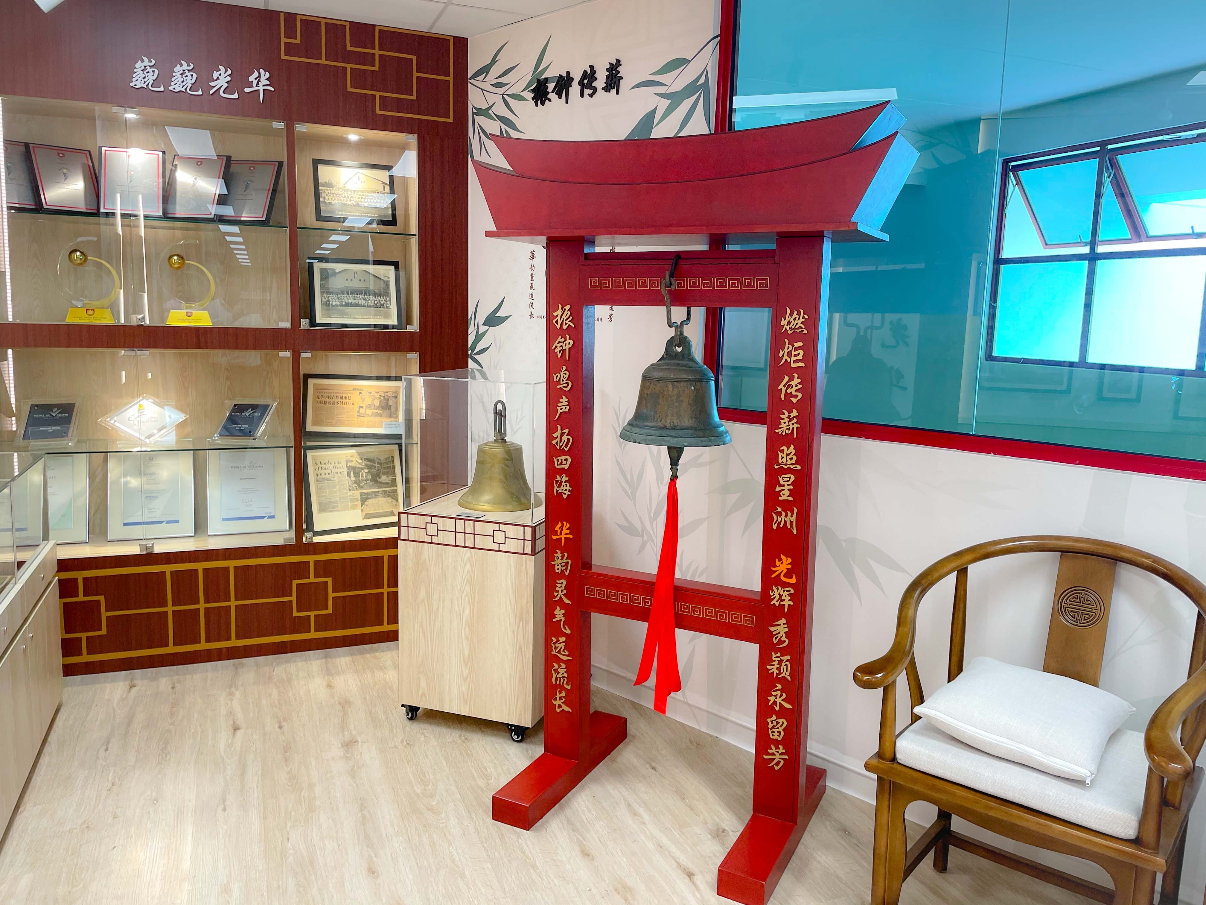 A red wooden bell stand with Chinese inscriptions holds two bronze bells, beside a wooden chair and glass display cabinets with awards.
