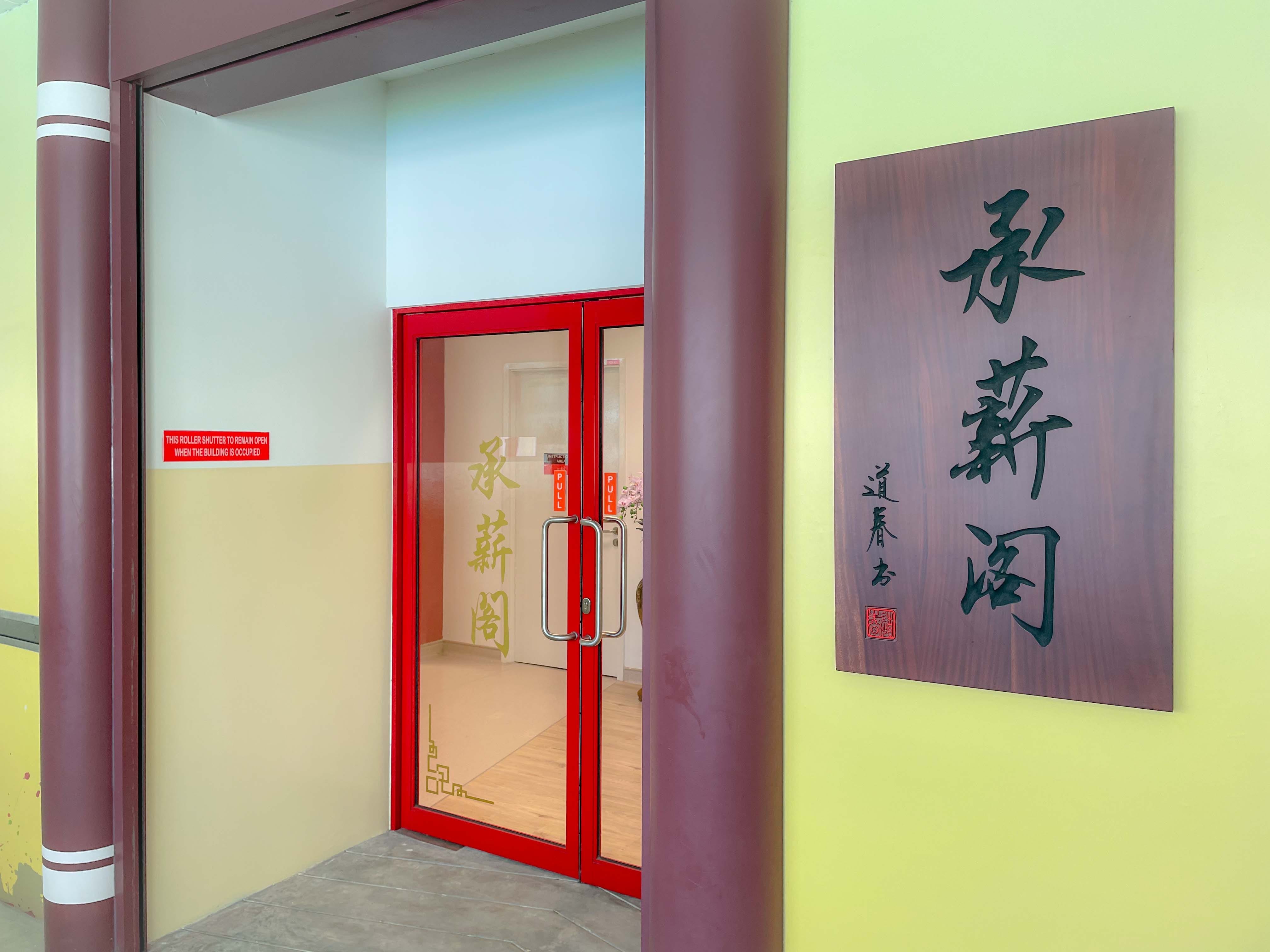 Entrance with red double doors and a wooden plaque displaying Chinese calligraphy characters on a yellow-green wall.