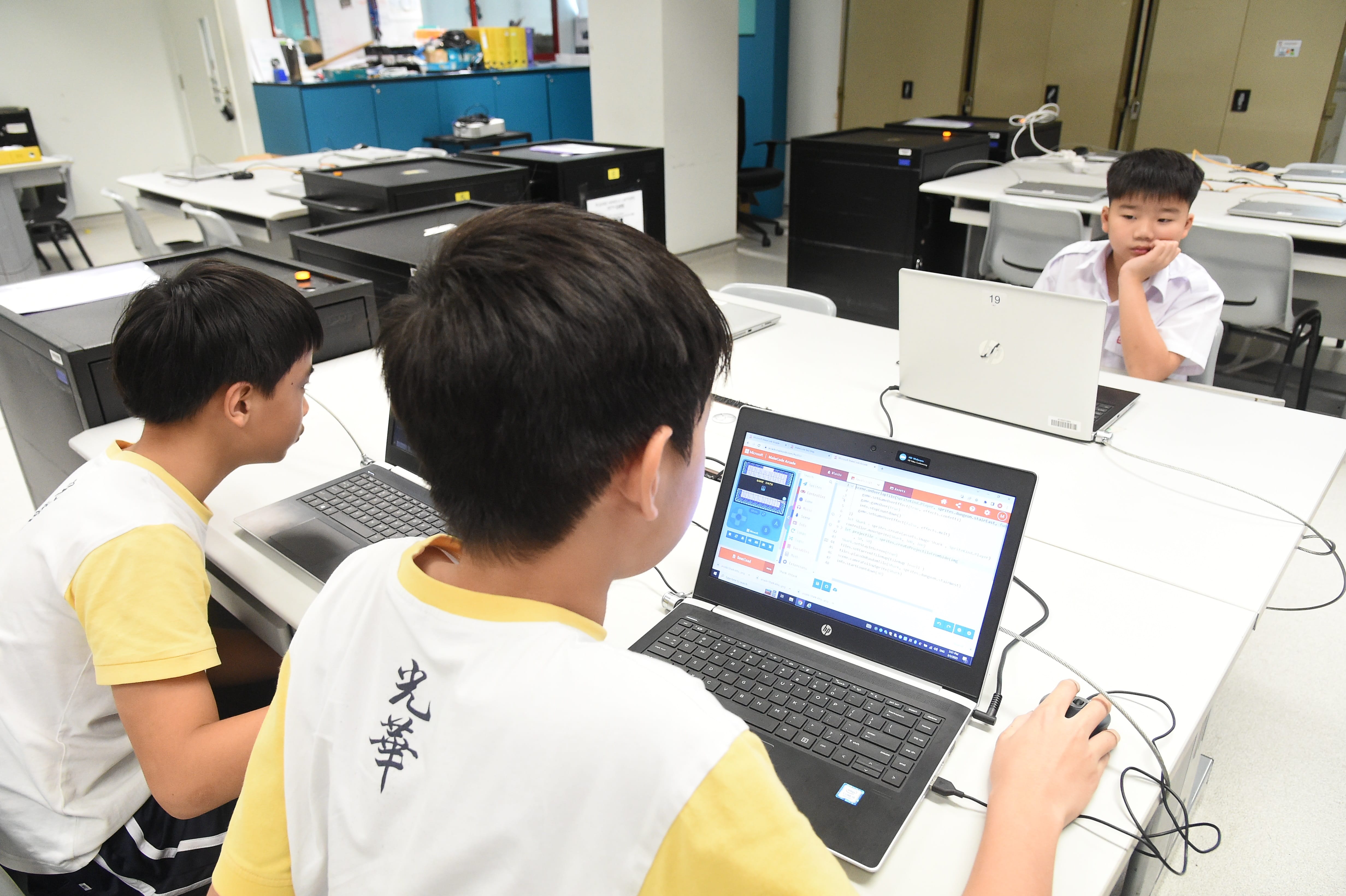 Three young boys in a school computer lab work on laptops, with one using a mouse to navigate a coding program.