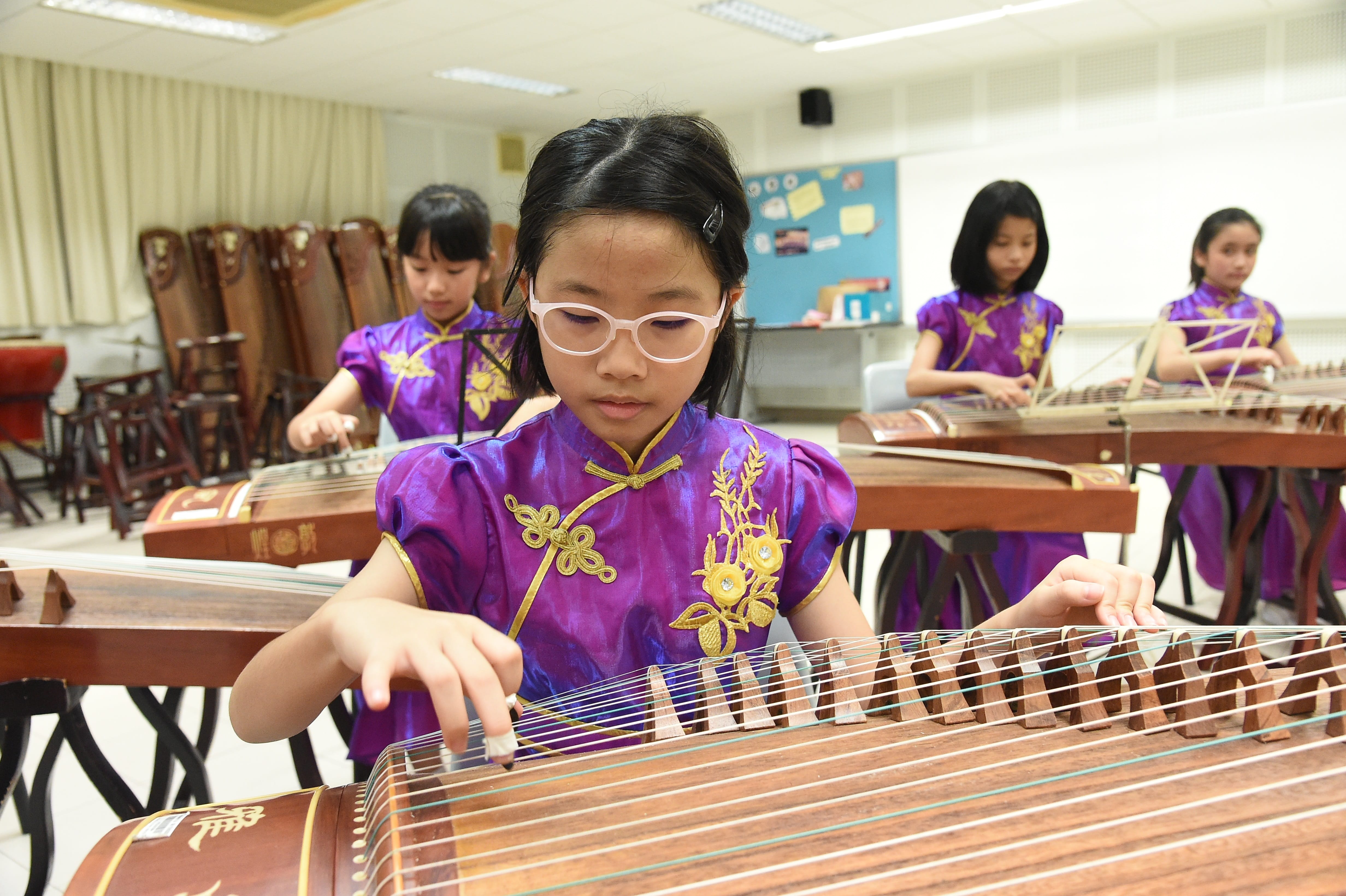 Four girls in purple embroidered qipao dresses play guzheng instruments in a classroom.