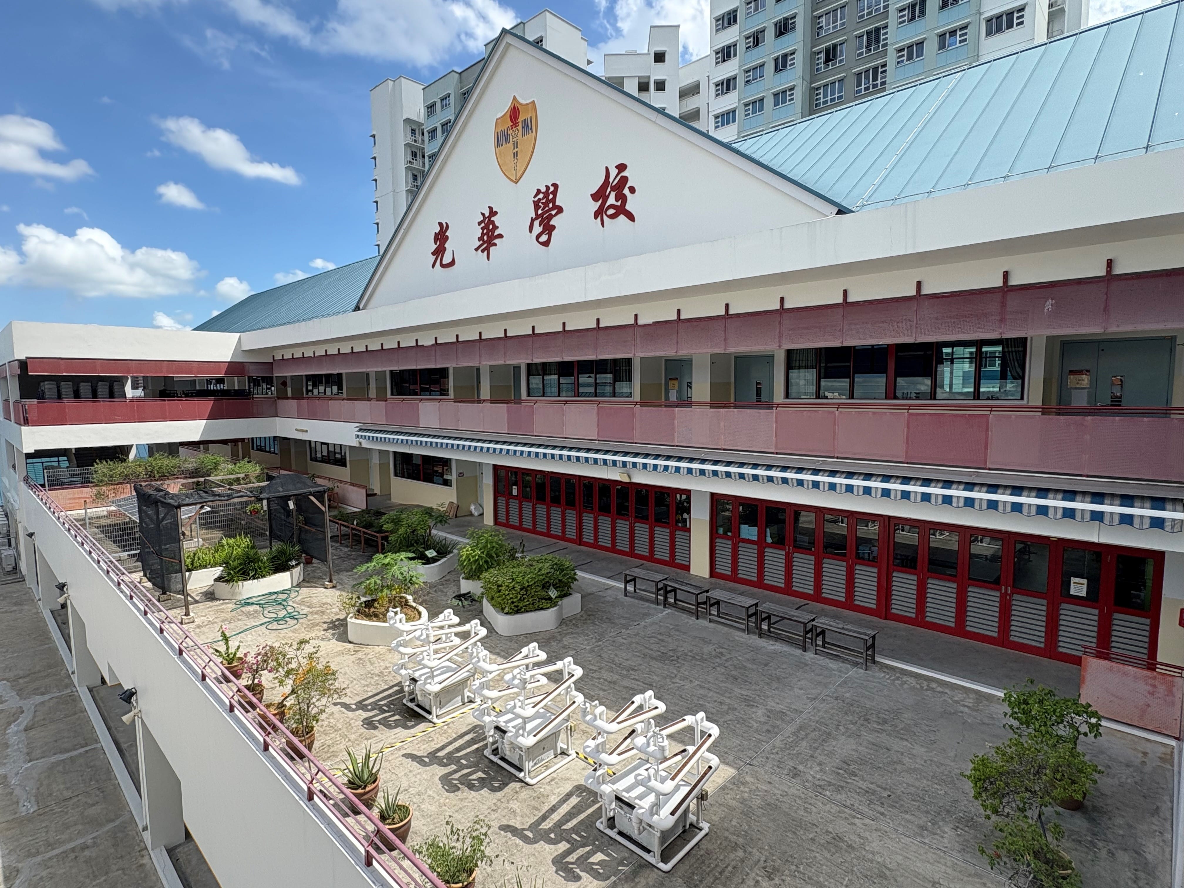 Exterior of Kong Hwa School with Chinese characters on a peaked roof, open courtyard, plants, and white pipe structures below.