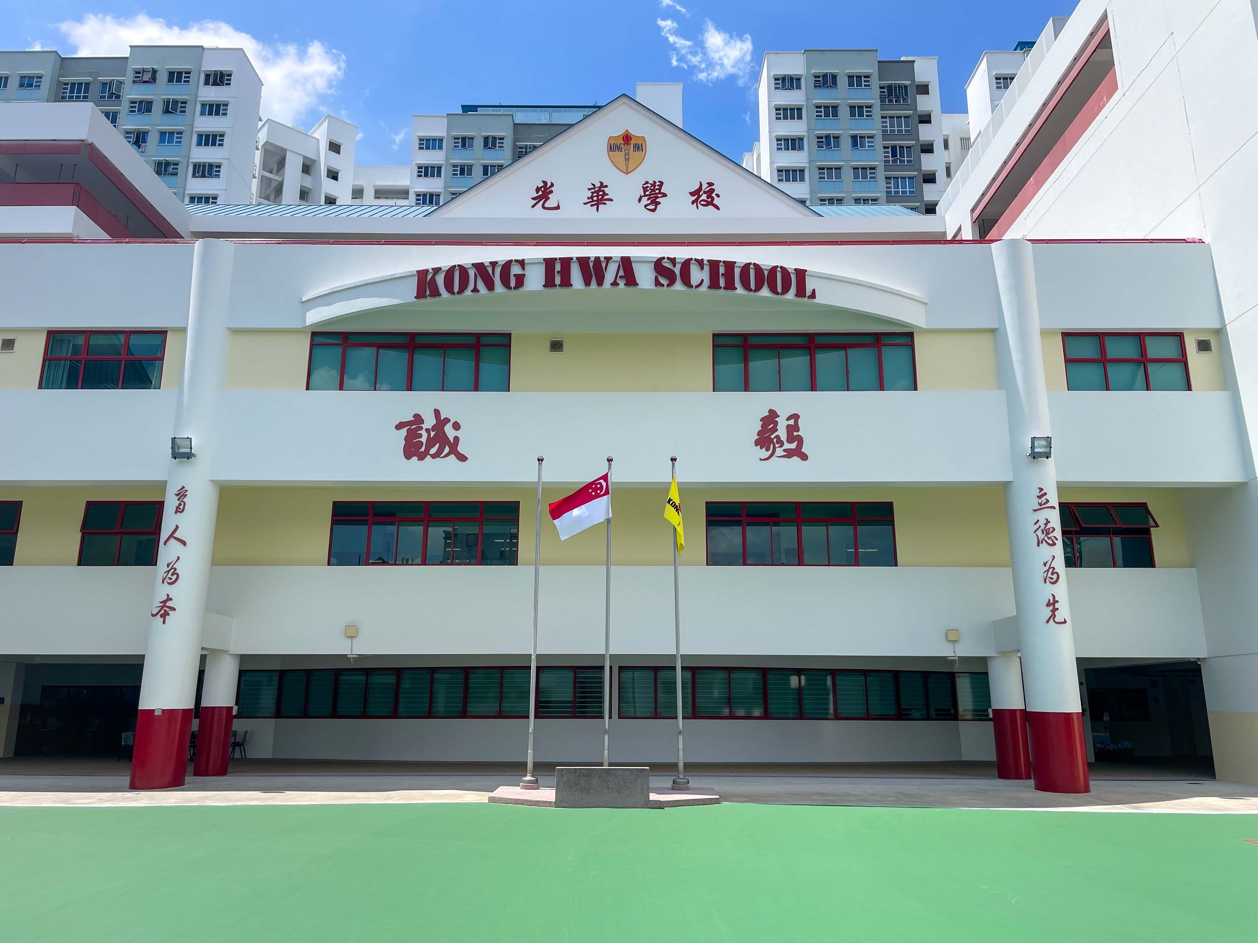 Front facade of Kong Hwa School with red and white walls, Chinese characters, and two flagpoles on a green courtyard.