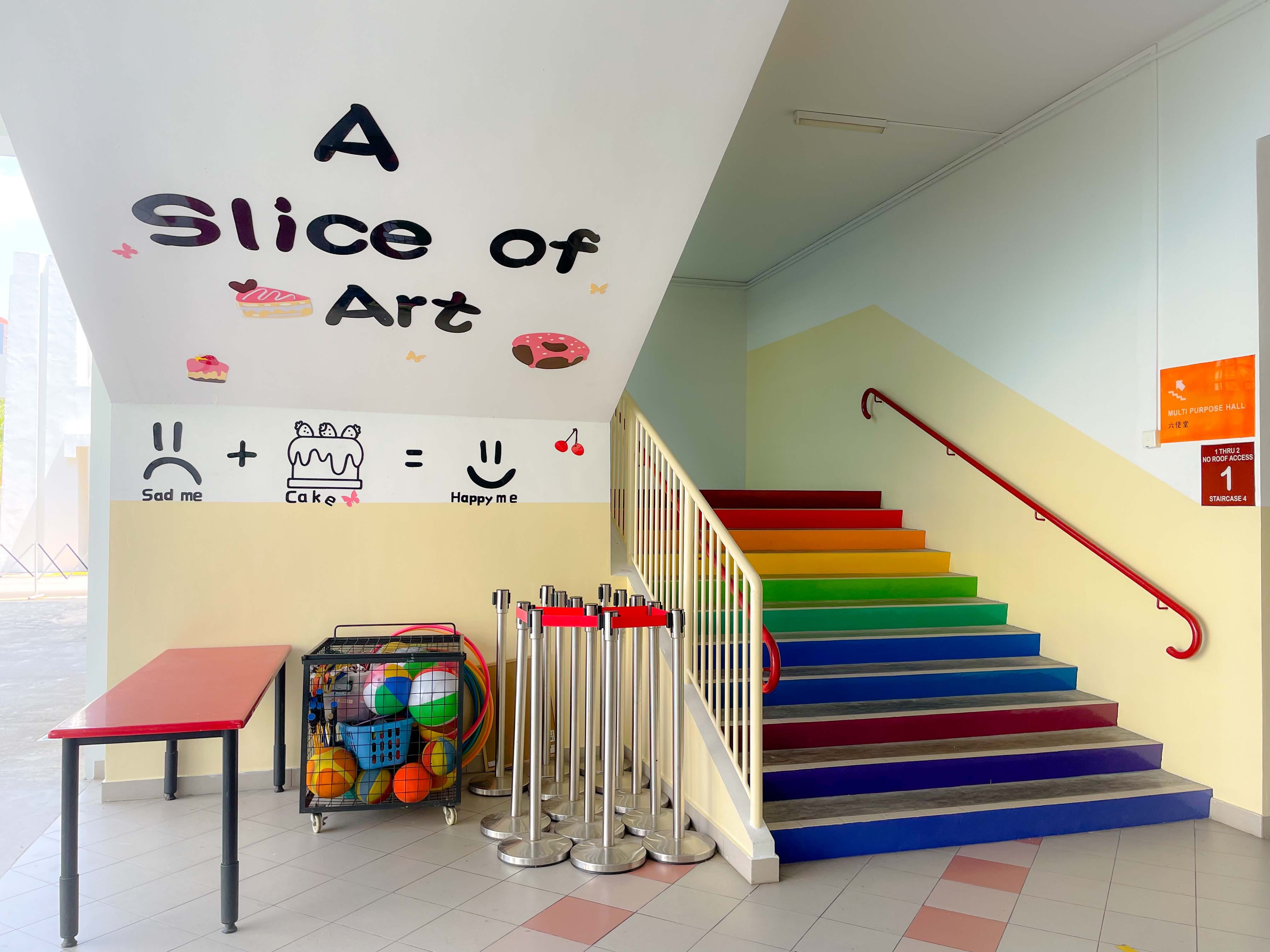 Rainbow-colored staircase beside a wall mural reading "A Slice of Art" with cake-themed drawings in a school hallway.