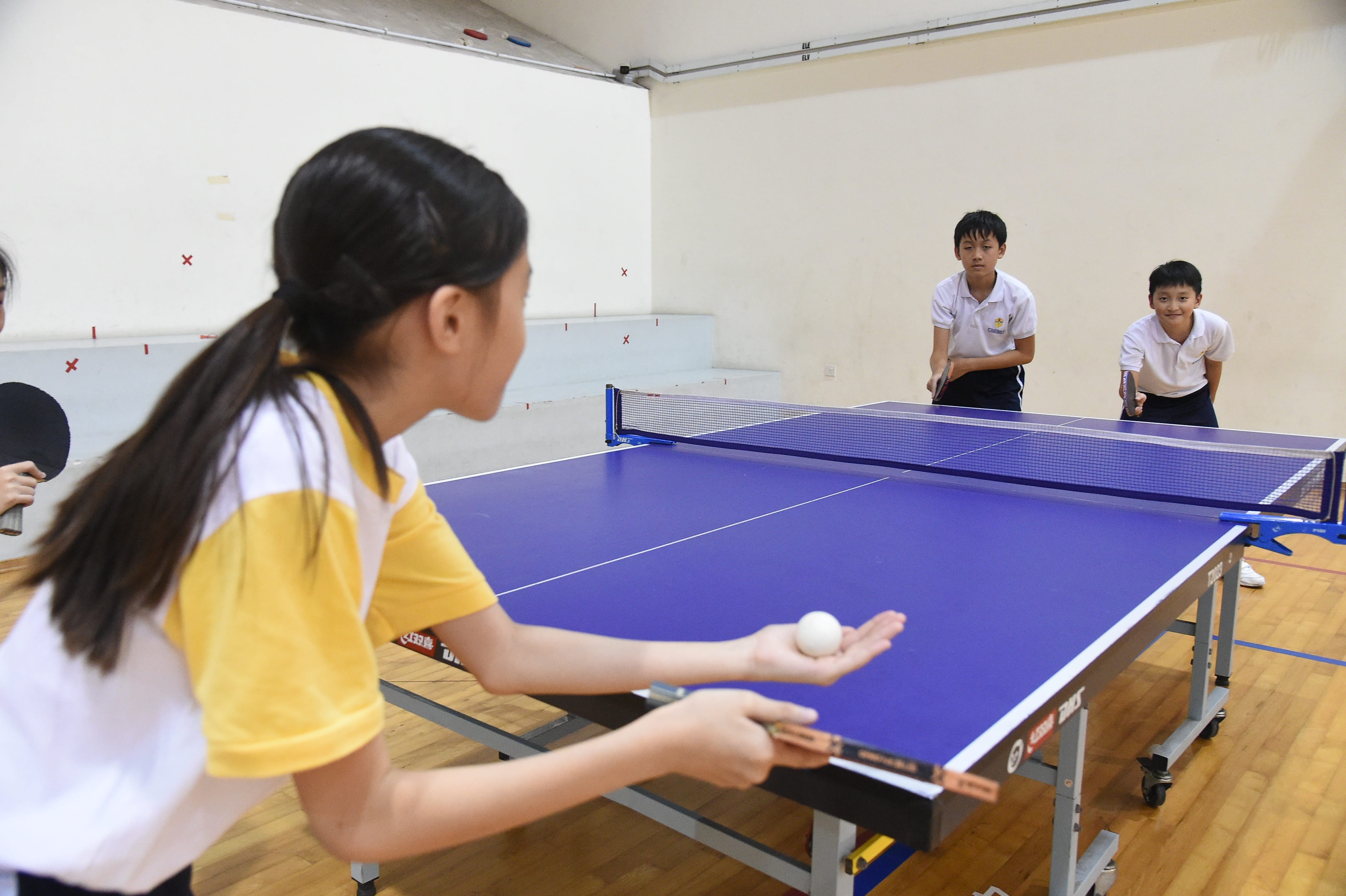 Four children play table tennis on a blue table indoors, with a girl preparing to serve the ball.