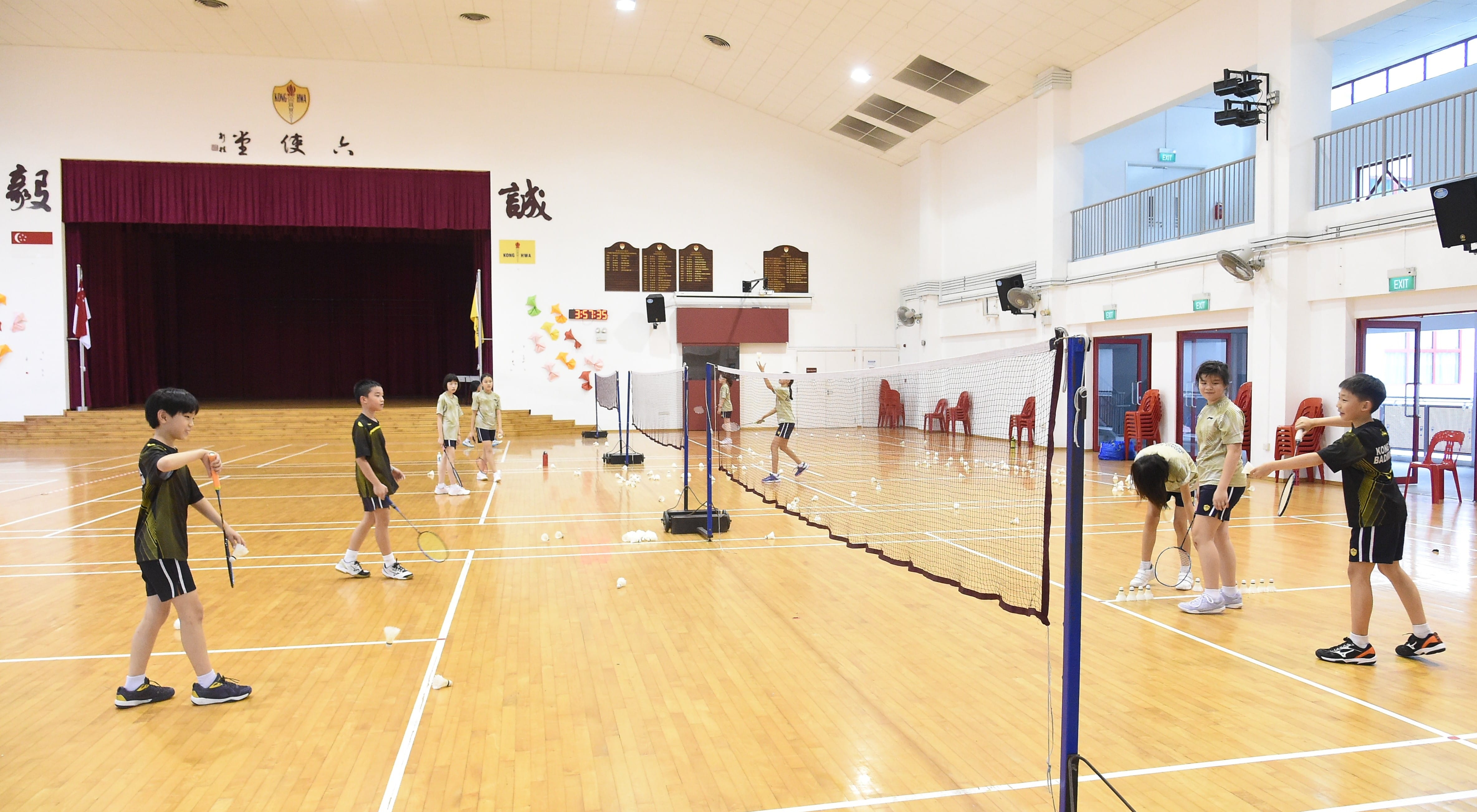 Several children practice badminton with rackets on an indoor wooden court with nets in a school hall.