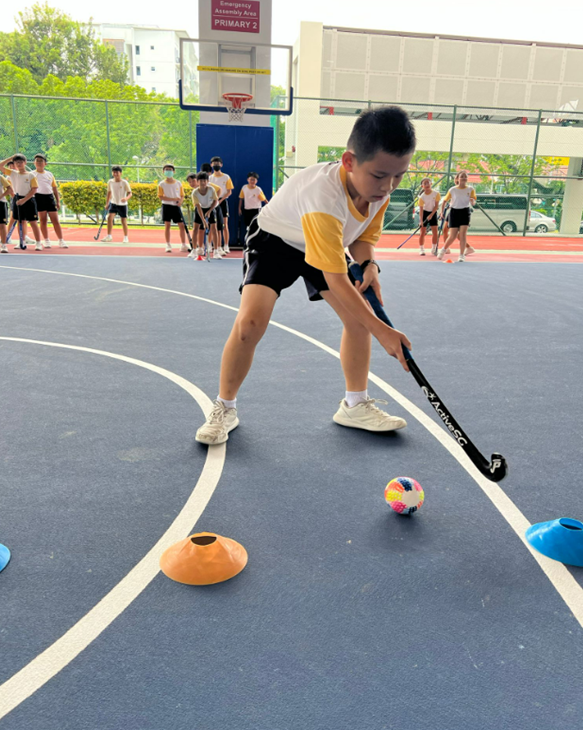 A P6 student practicing dribbling a ball with a hockey stick
