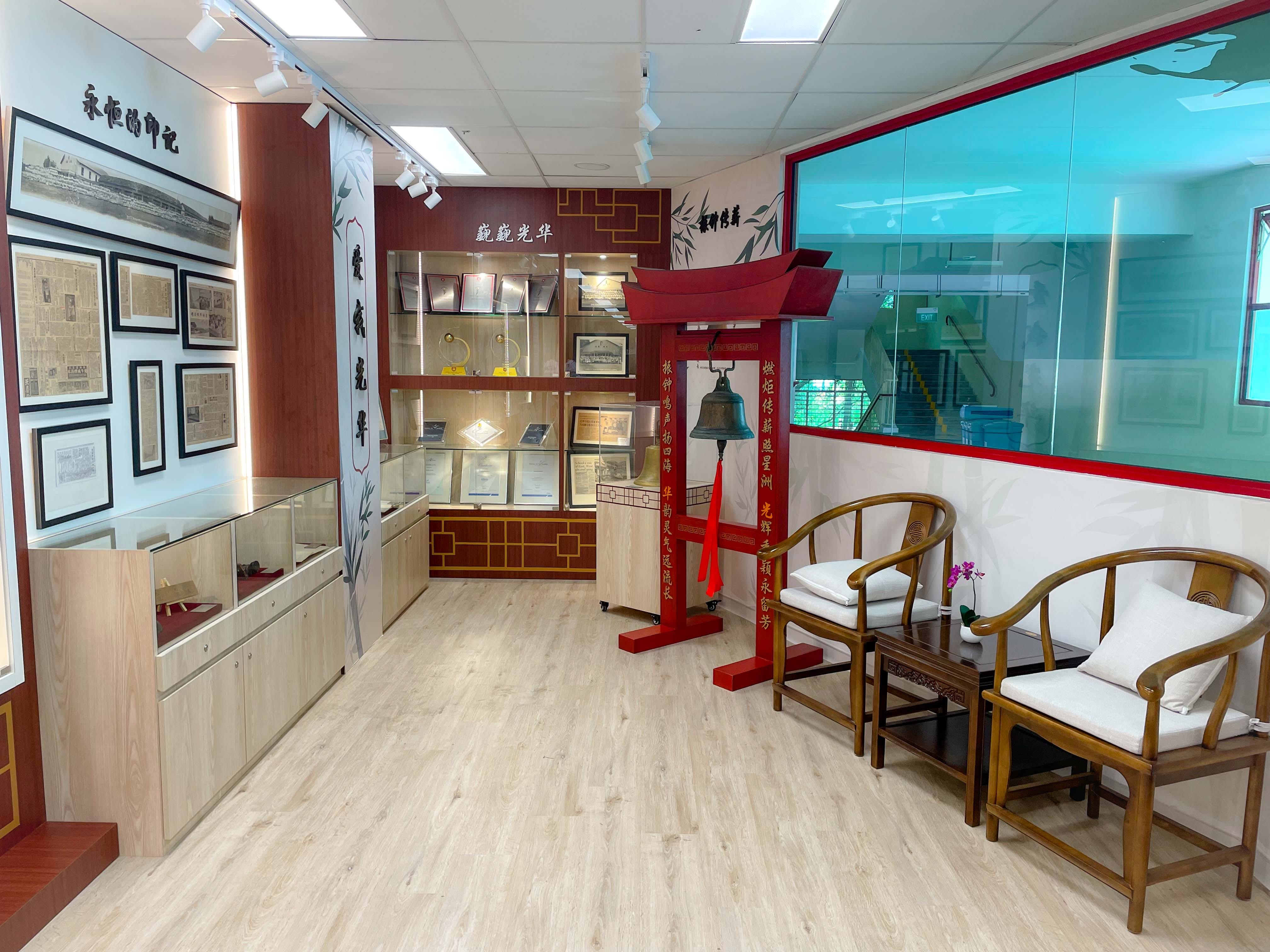 A Chinese heritage room with a red bell stand, display cases, framed documents on walls, and traditional wooden chairs.