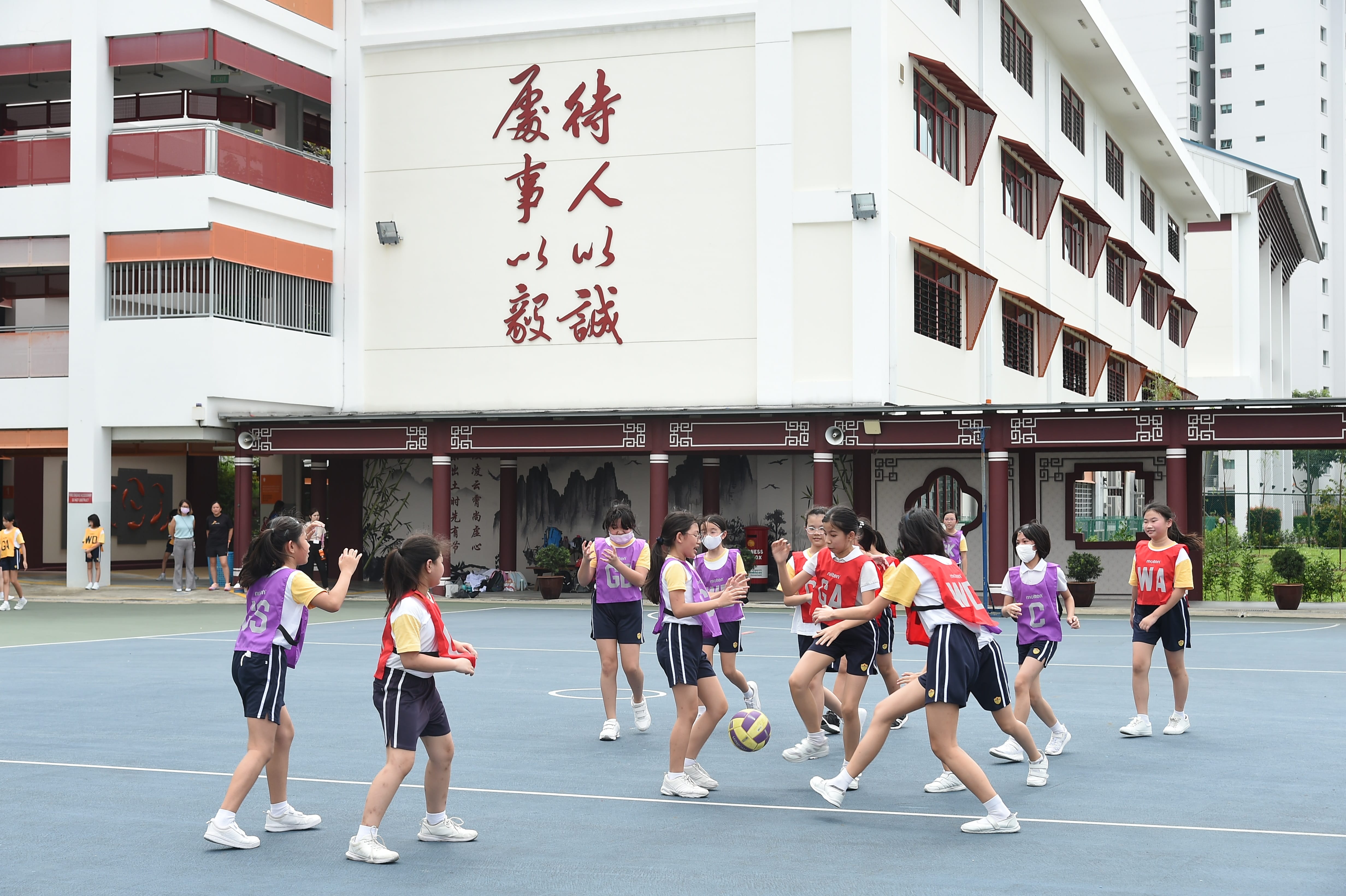 Girls in red and purple bibs play netball on an outdoor court.