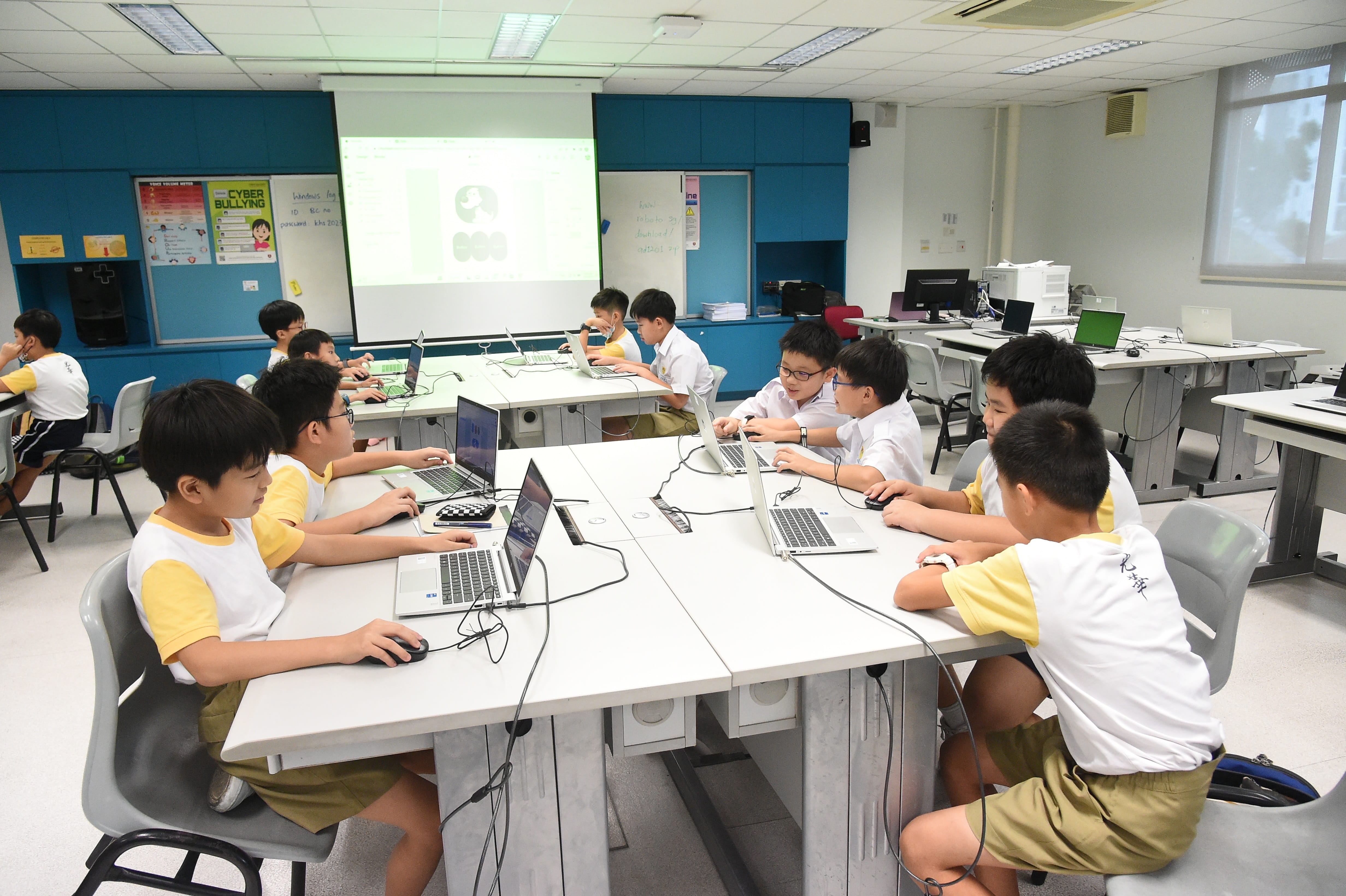 Students in uniforms sit at tables using laptops in a computer classroom with a projected screen at the front.