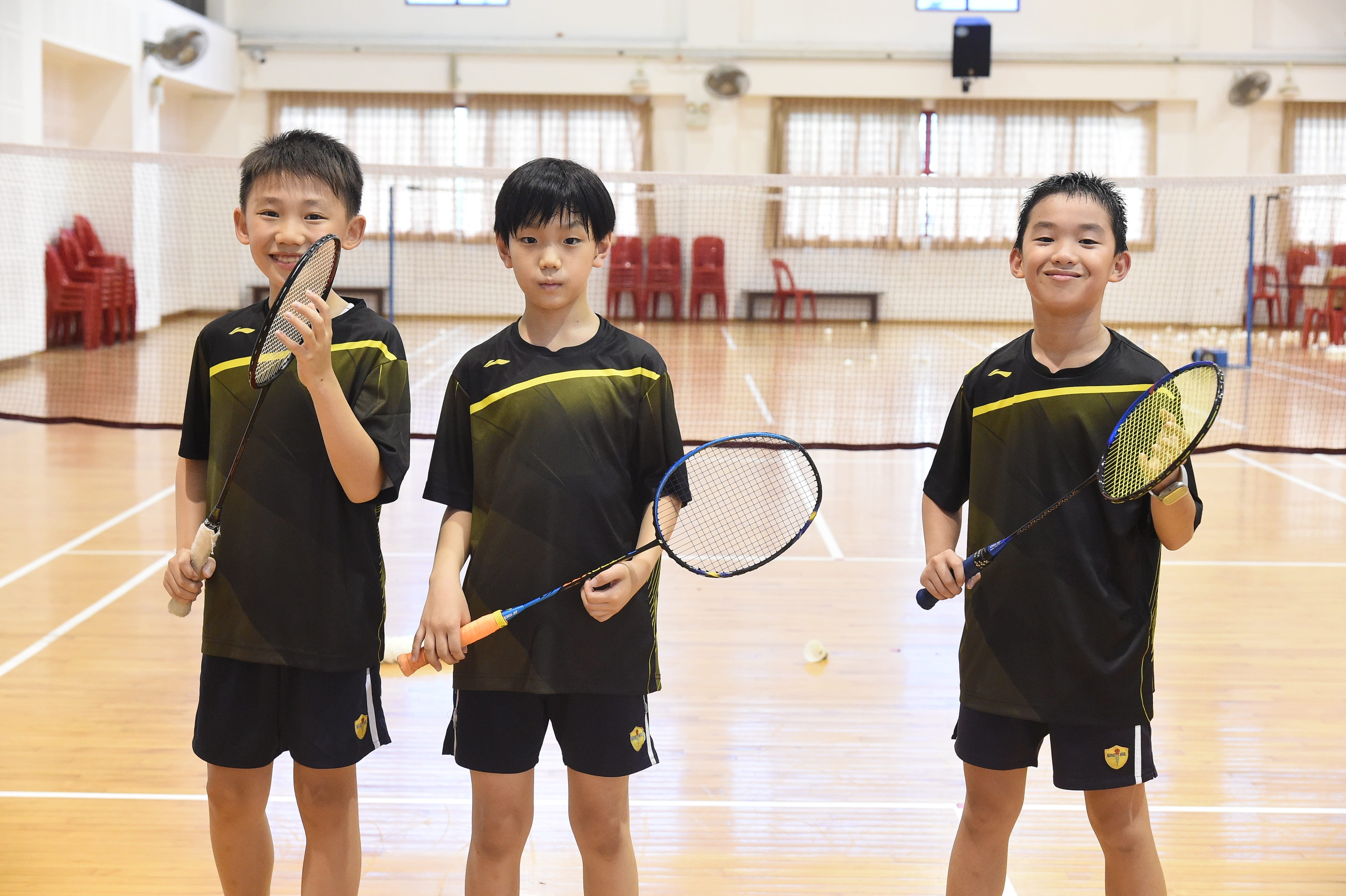 Three boys in black and yellow uniforms hold badminton rackets and smile inside an indoor badminton court.