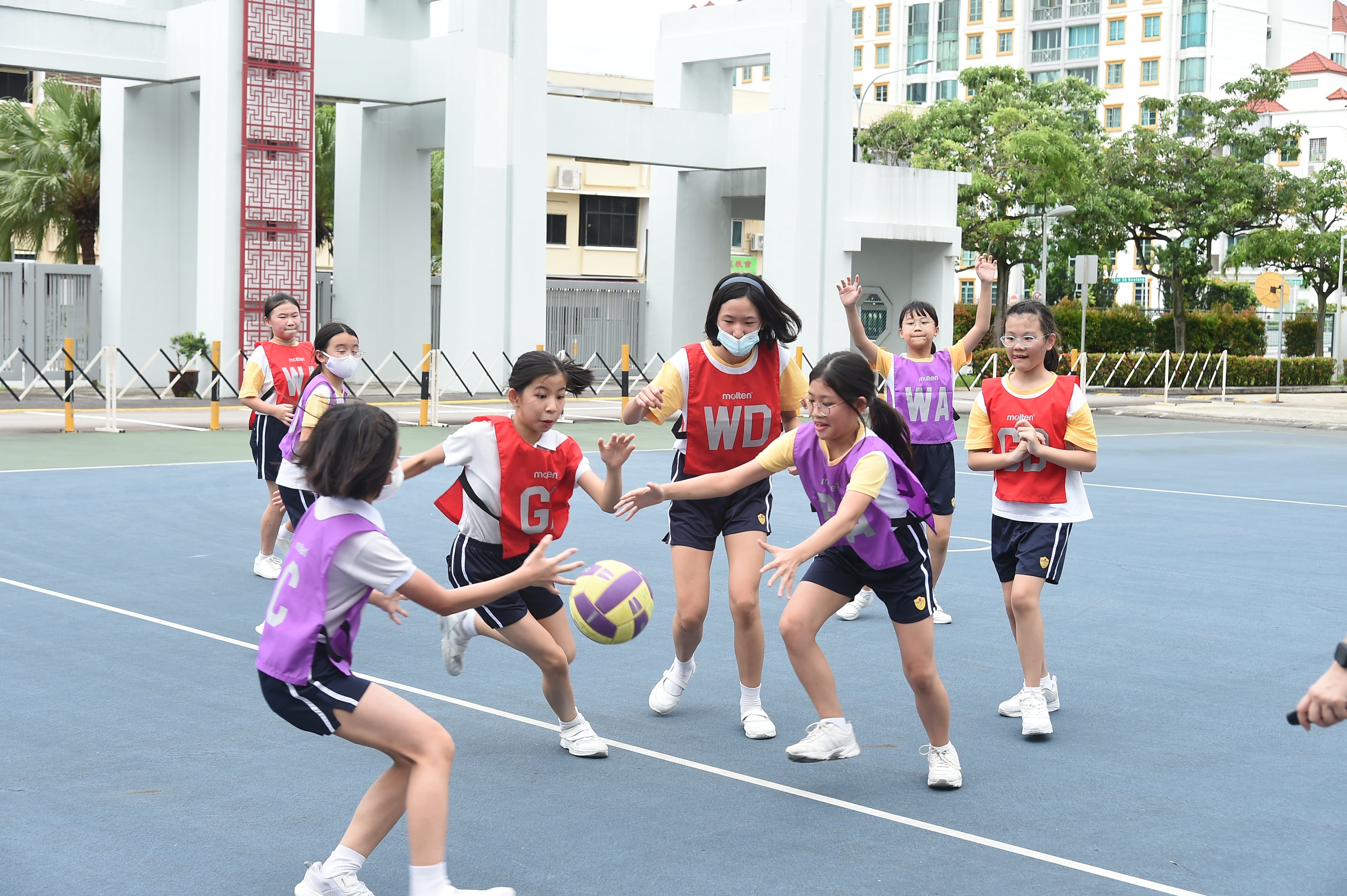 Girls in red and purple bibs play netball on an outdoor court, competing for a yellow and purple ball.