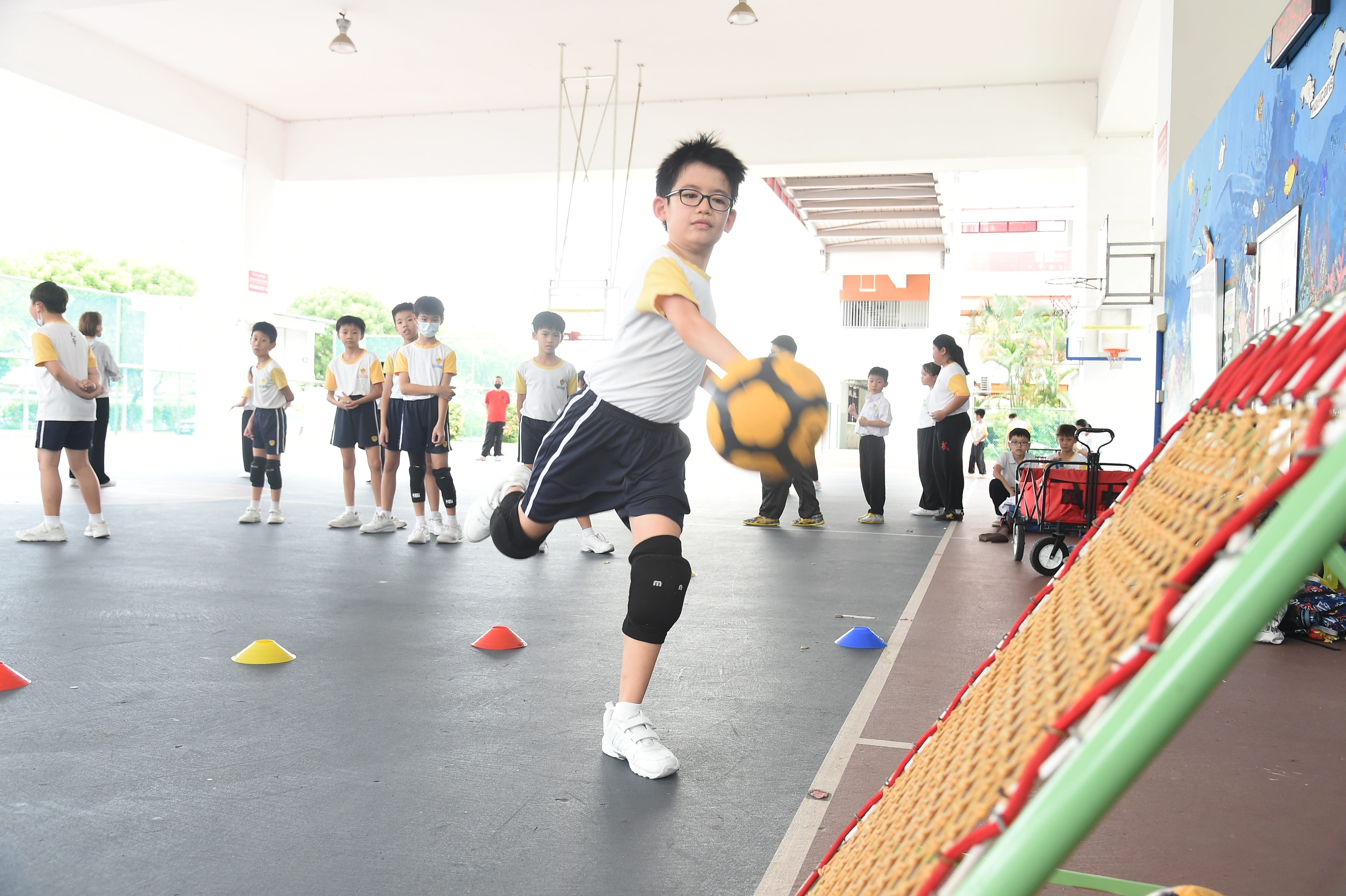 Boy with glasses throwing a yellow soccer ball in a school covered court while classmates watch in the background.