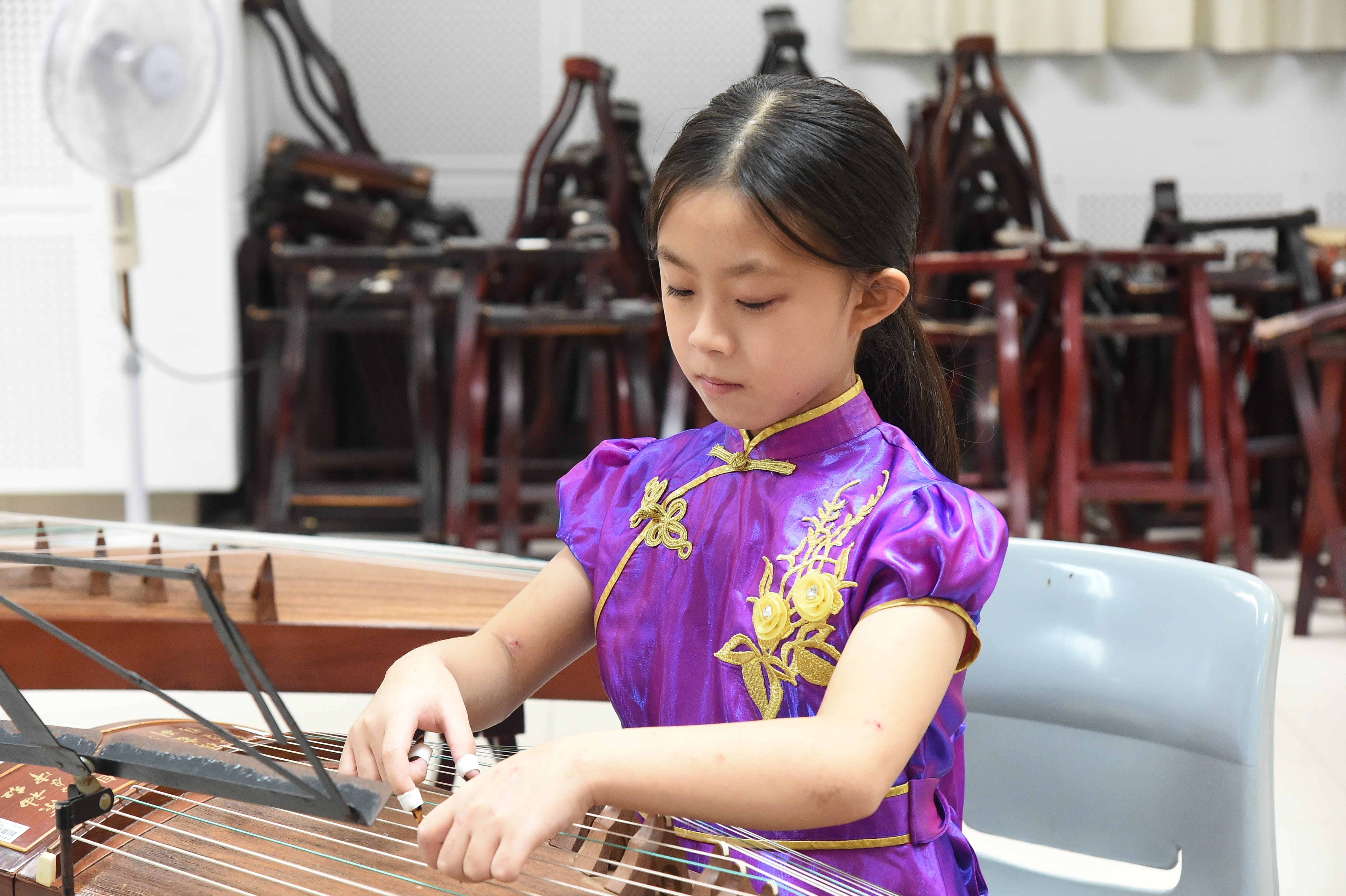 Young girl in a purple traditional Chinese dress playing a guzheng in a music classroom.