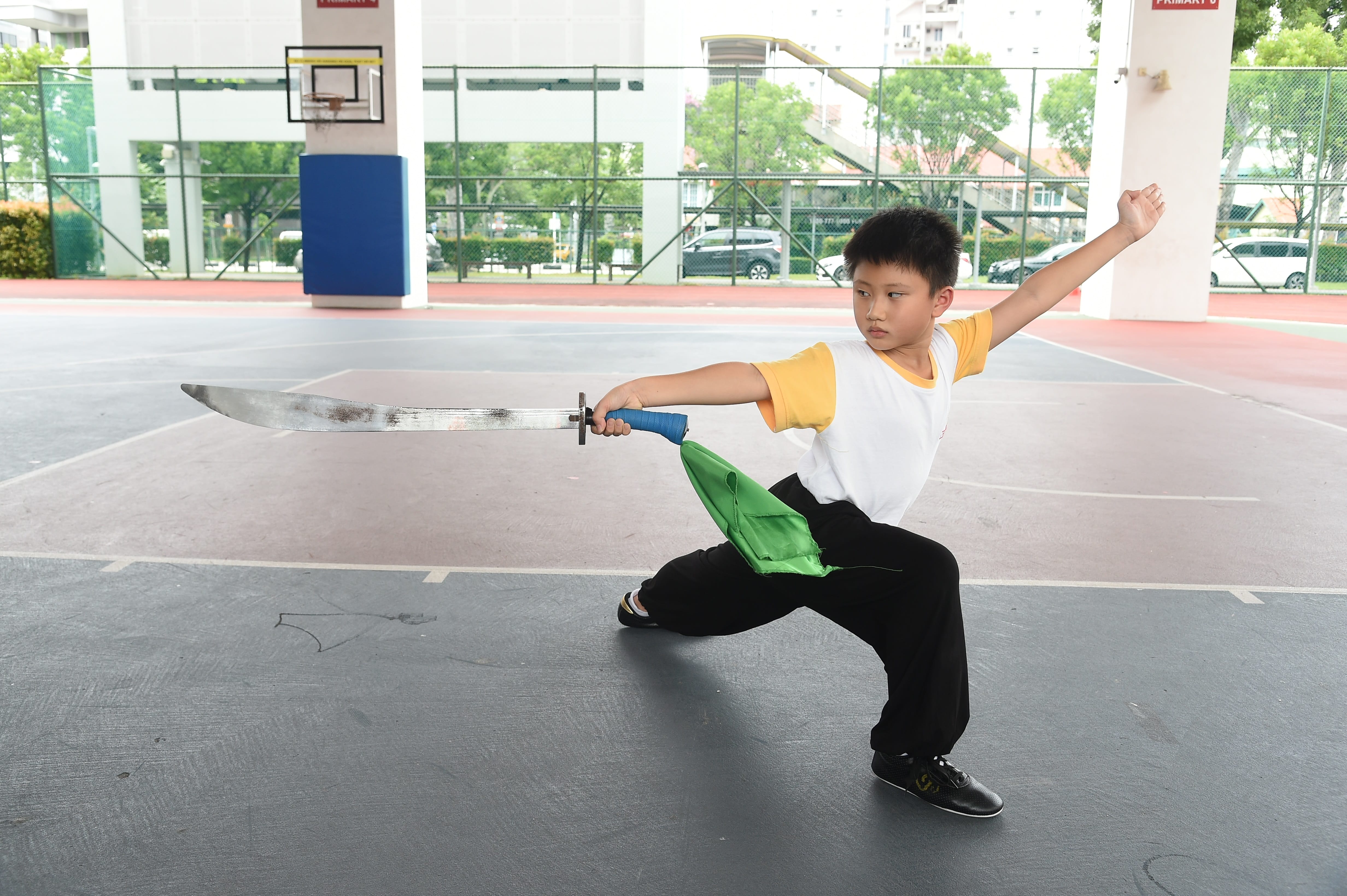 A young boy in a wide stance holds a large sword with a blue handle while practicing wushu on an outdoor court.