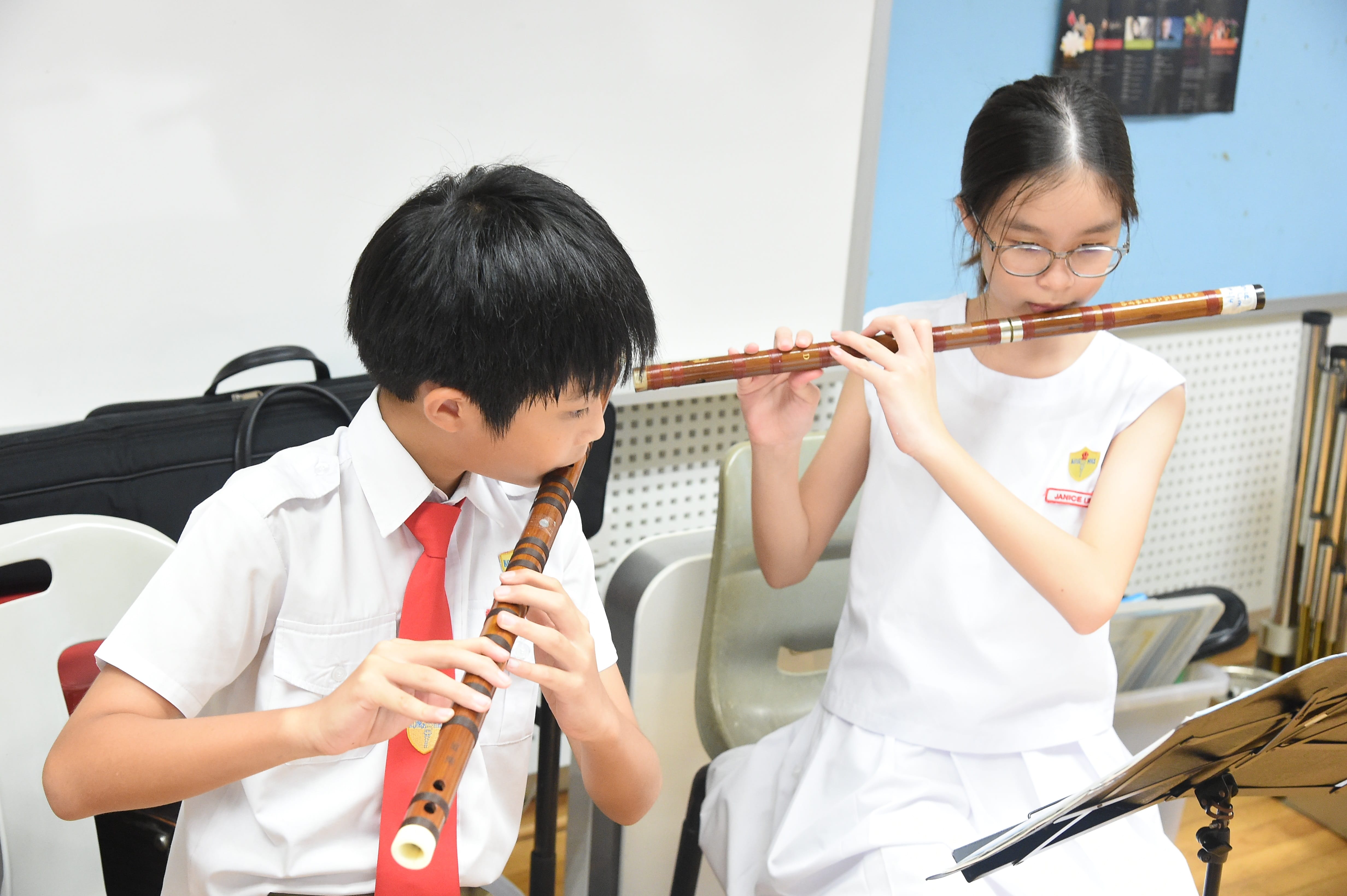 Two young students in school uniforms play wooden flutes in a classroom setting.