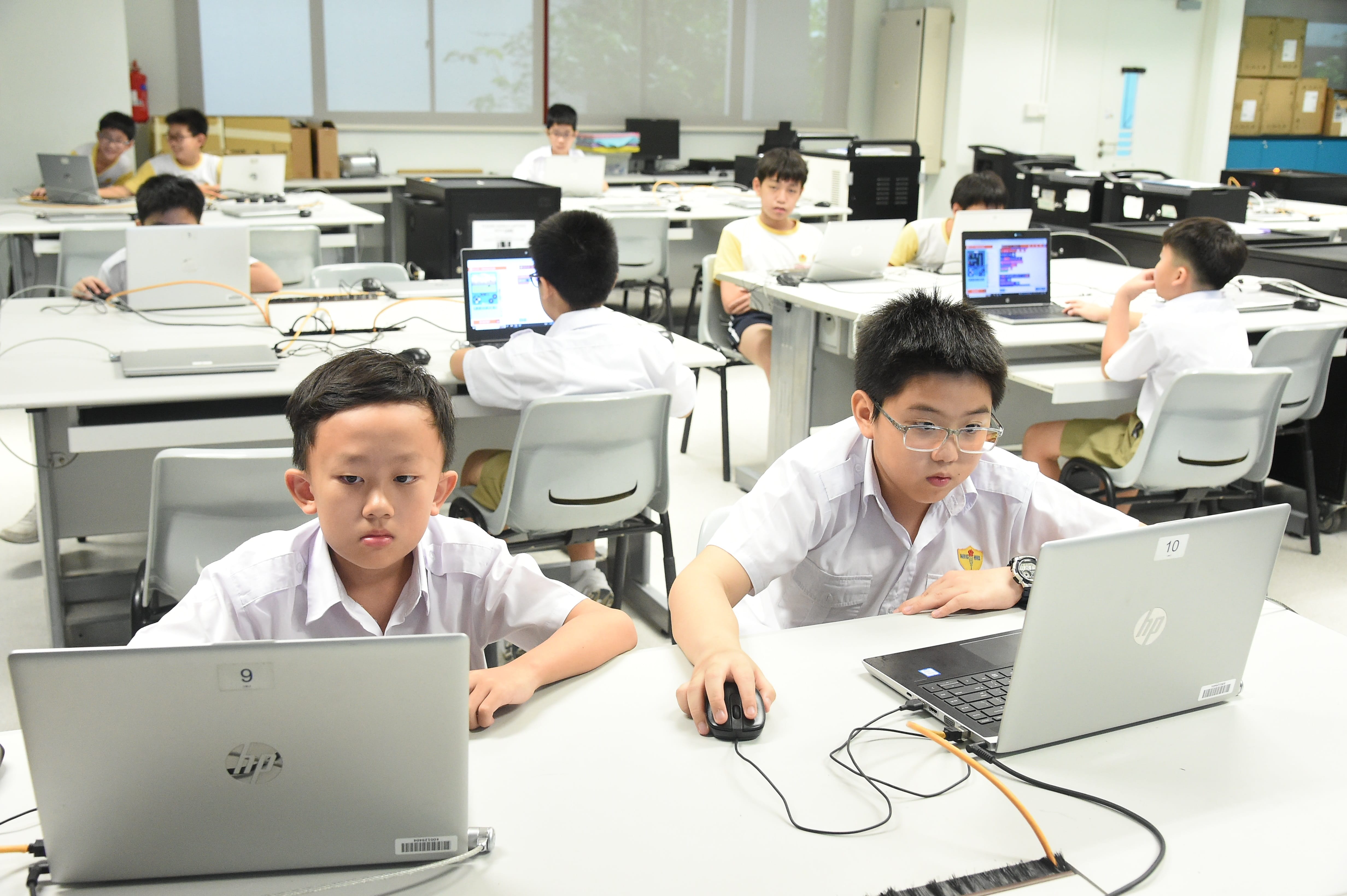 Several school boys in white uniforms work on laptops in a bright computer classroom.