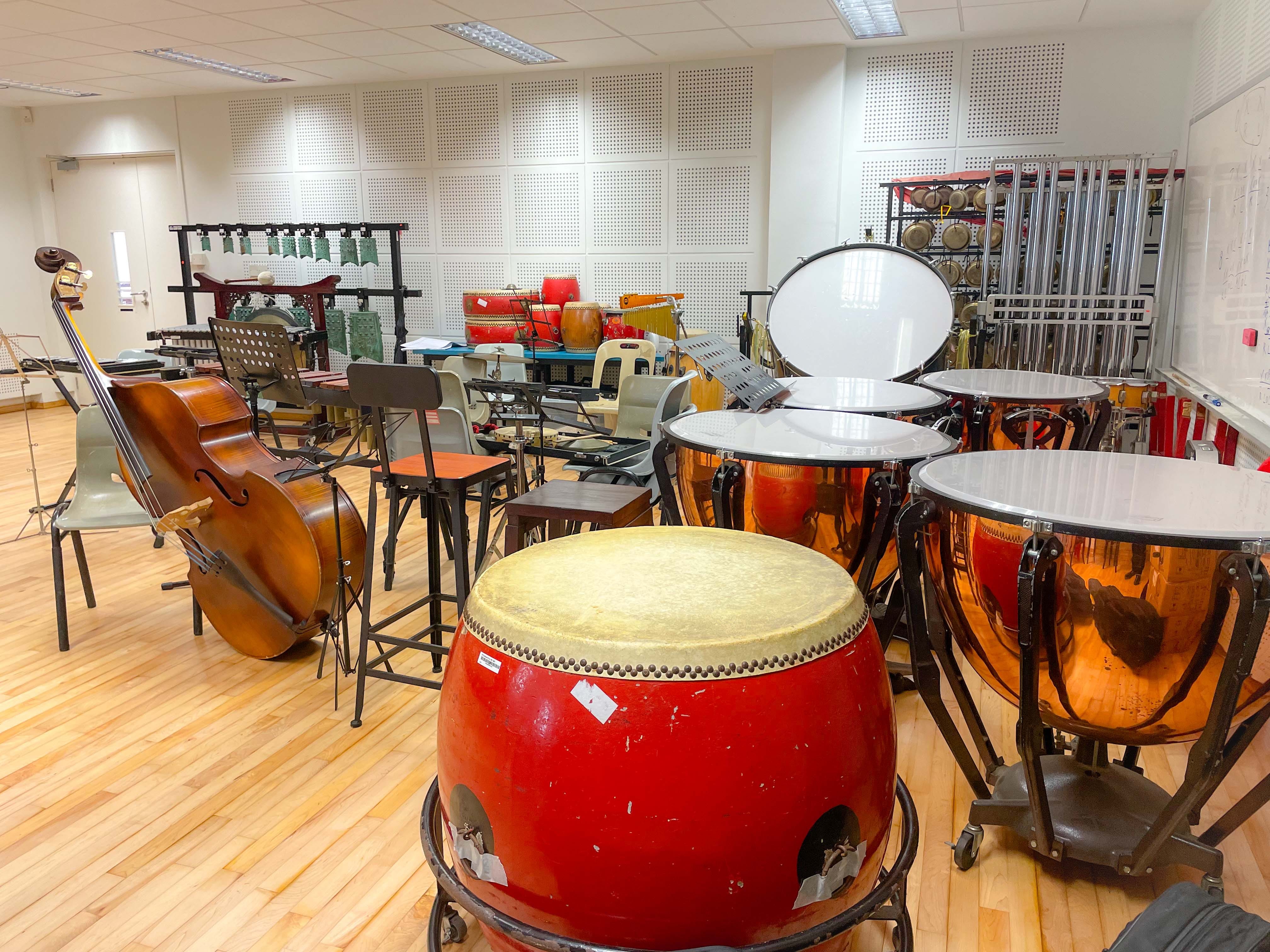 A music room with timpani drums, a red taiko drum, a double bass, bells, and various percussion instruments on a wood floor.