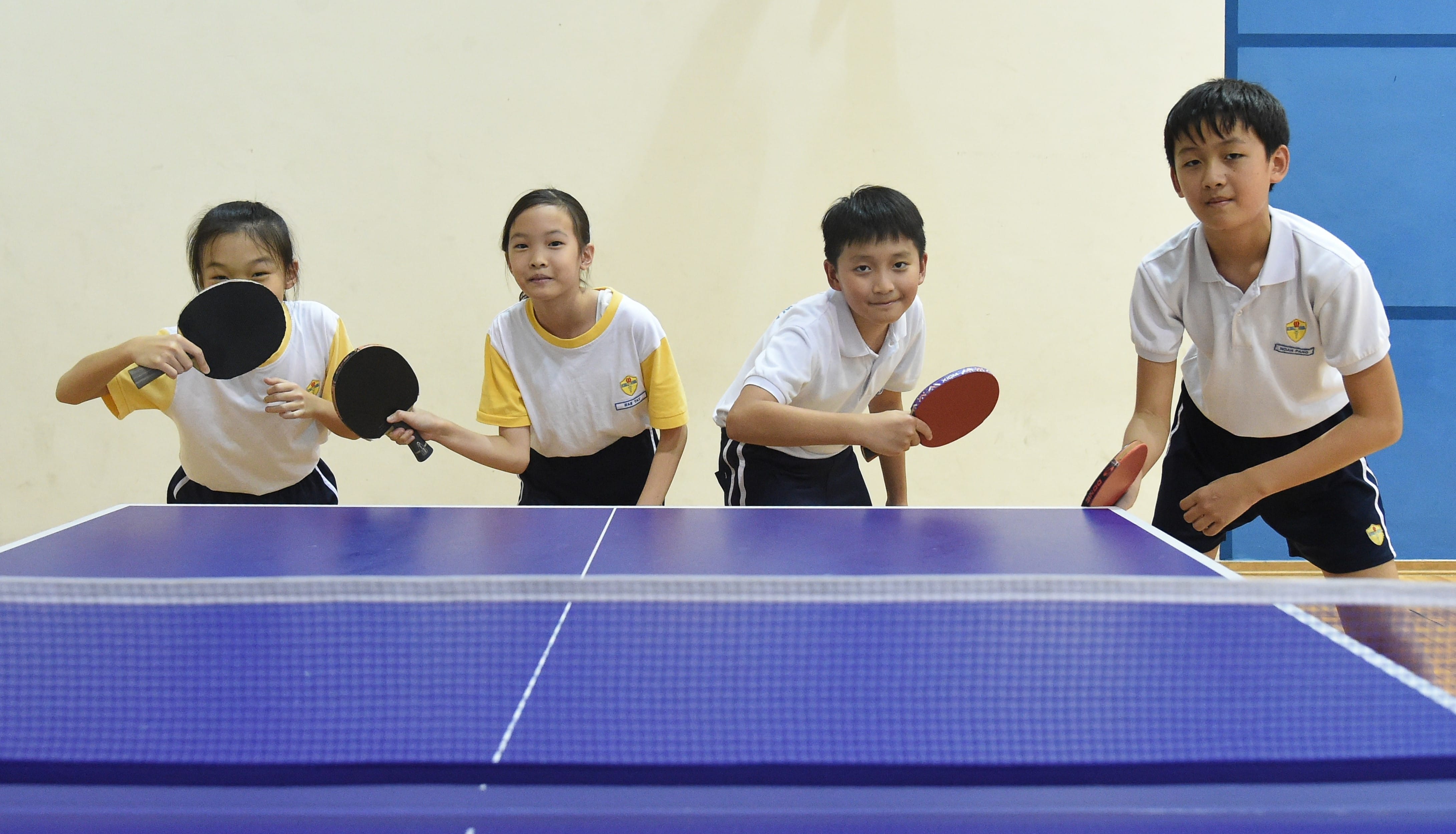 Four school children in uniforms hold table tennis paddles behind a blue ping-pong table indoors.