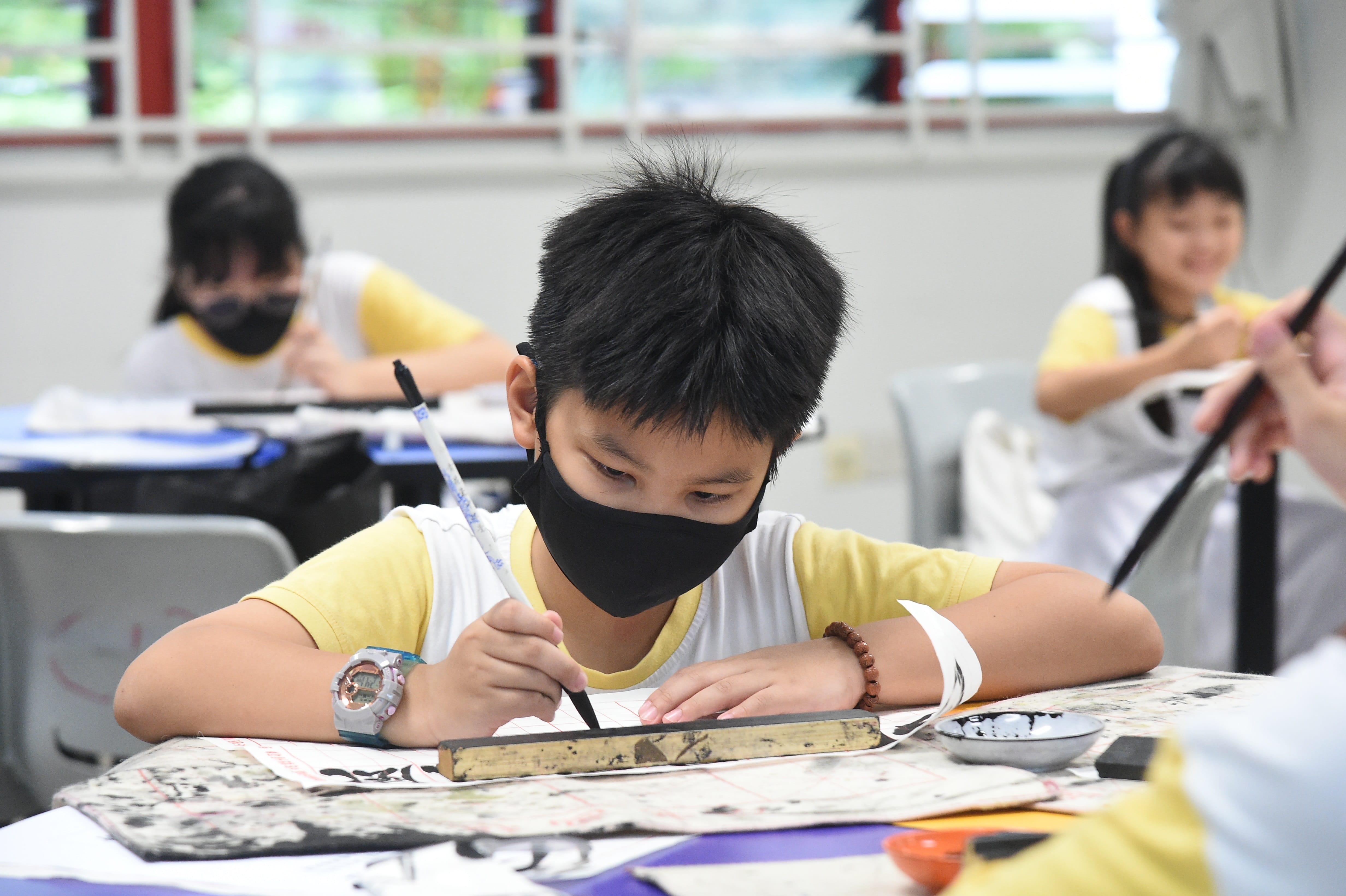 A masked boy practices calligraphy with a brush at a desk, with two other masked children in the background.