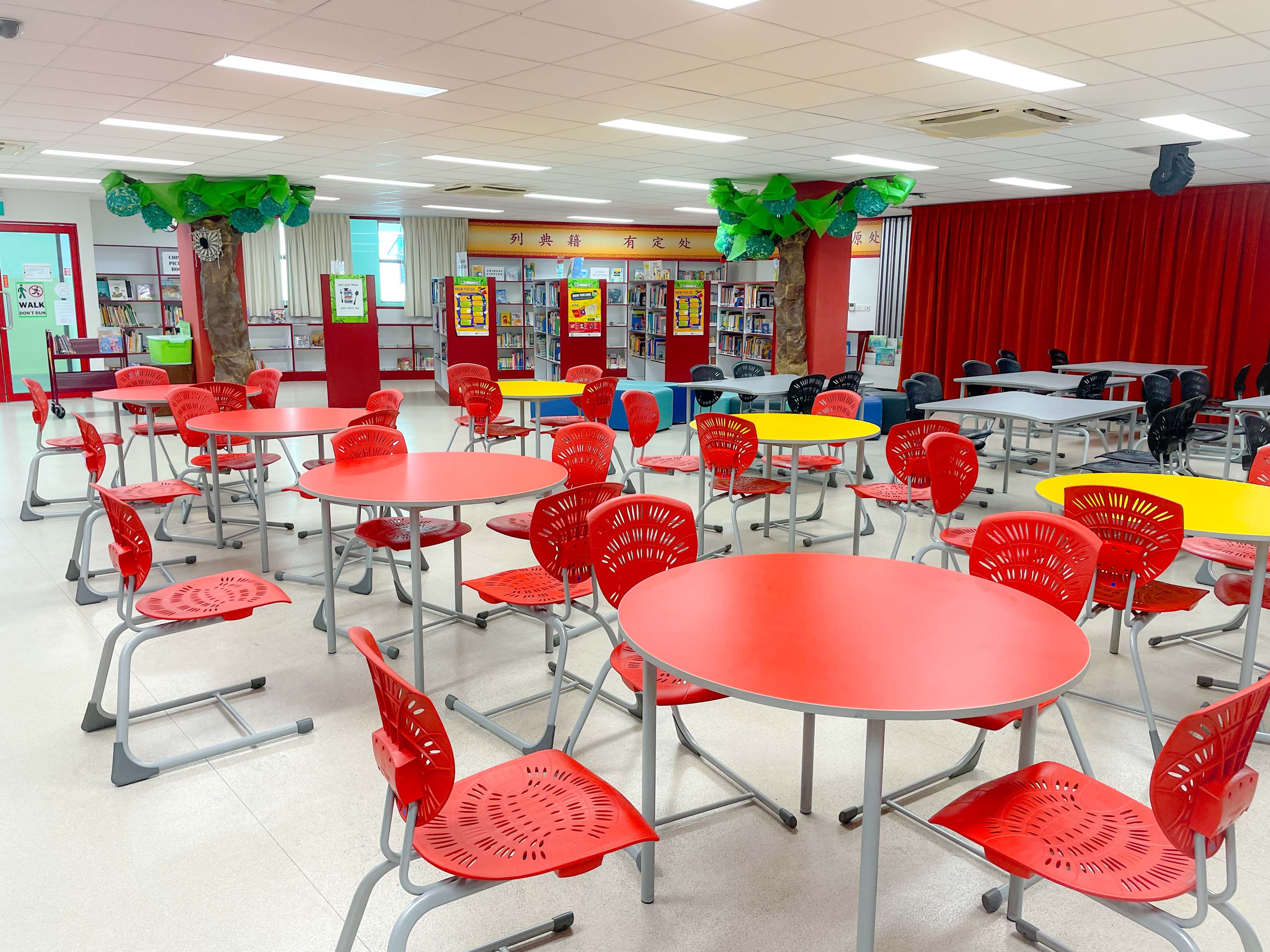 A school library with red and yellow round tables, red plastic chairs, bookshelves, and decorative tree columns.