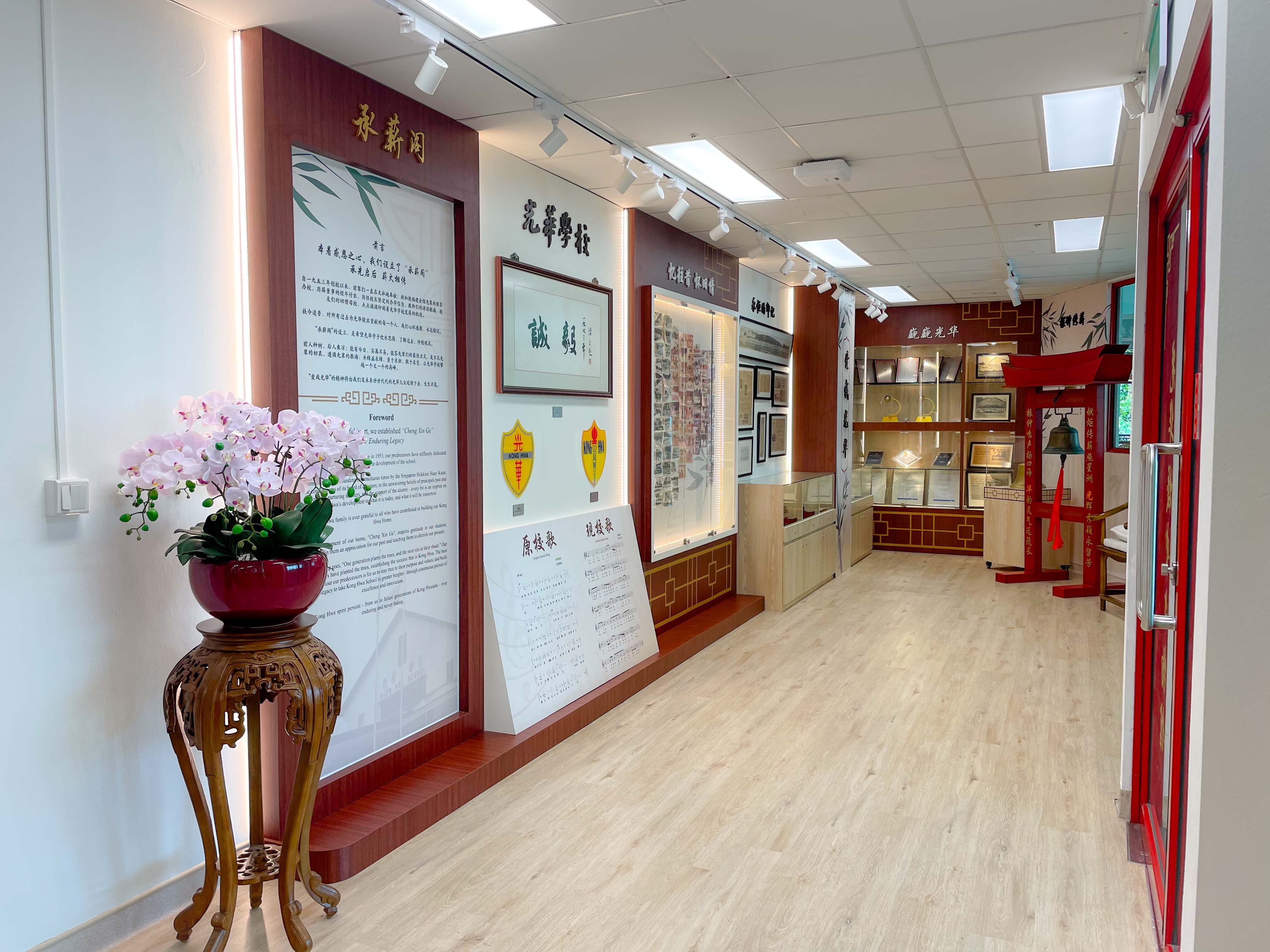 A Chinese heritage museum corridor with red wood panels, framed displays, a school bell, and pink orchids on a carved stand.