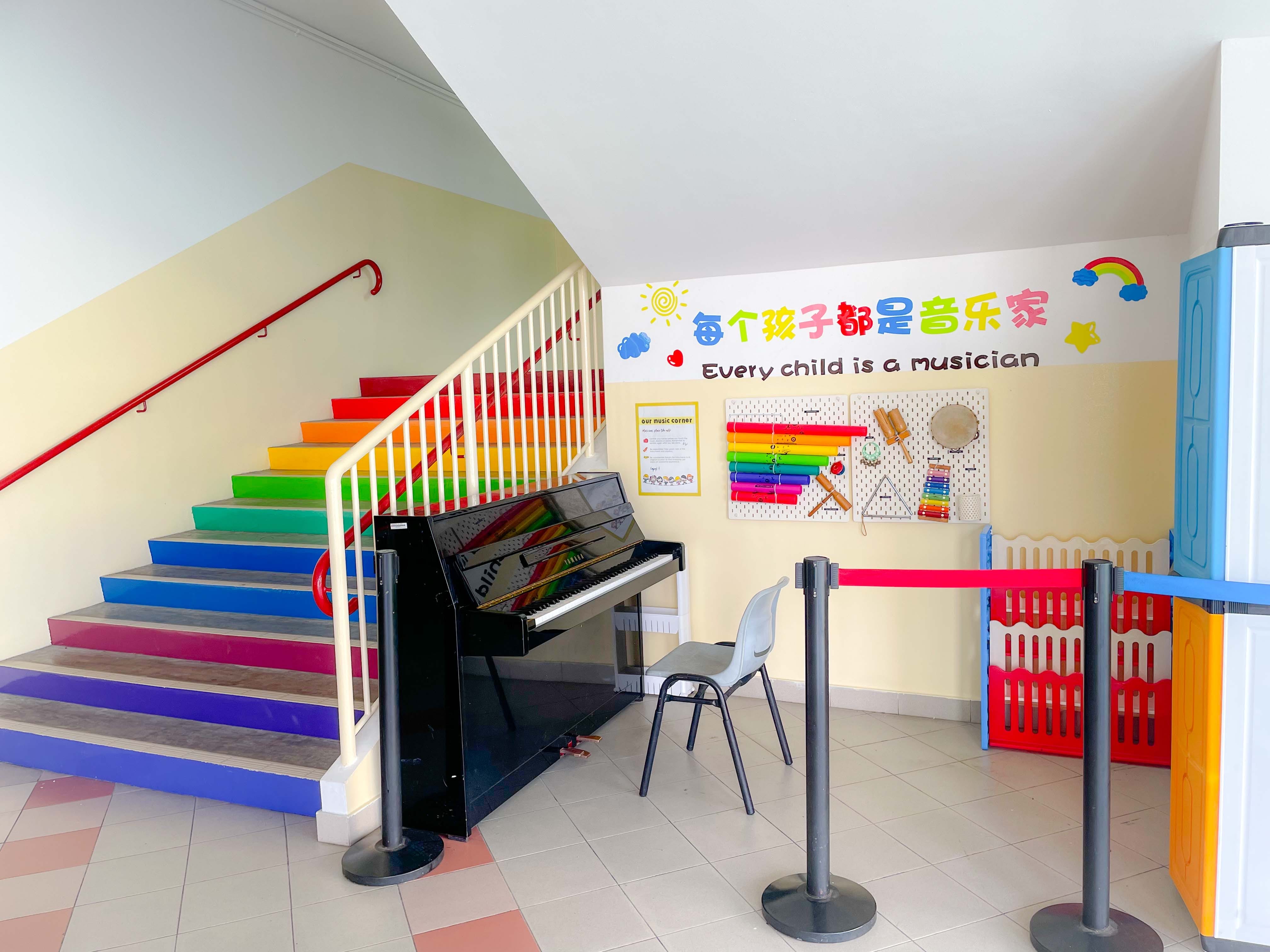 Colorful rainbow staircase beside a black upright piano, with a wall reading "Every child is a musician" and children's instruments displayed.