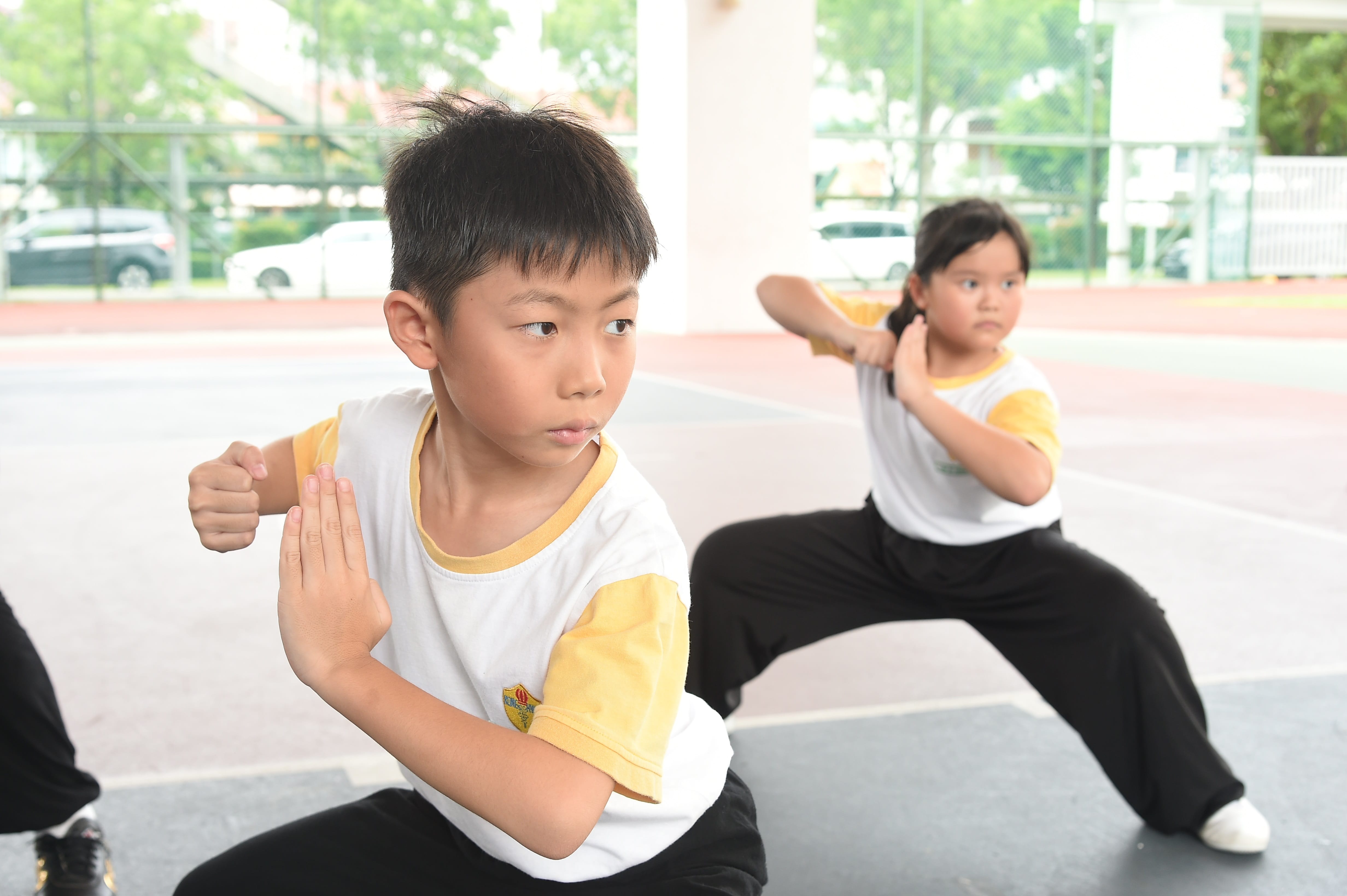 Two children in white and yellow uniforms practice martial arts stances outdoors.
