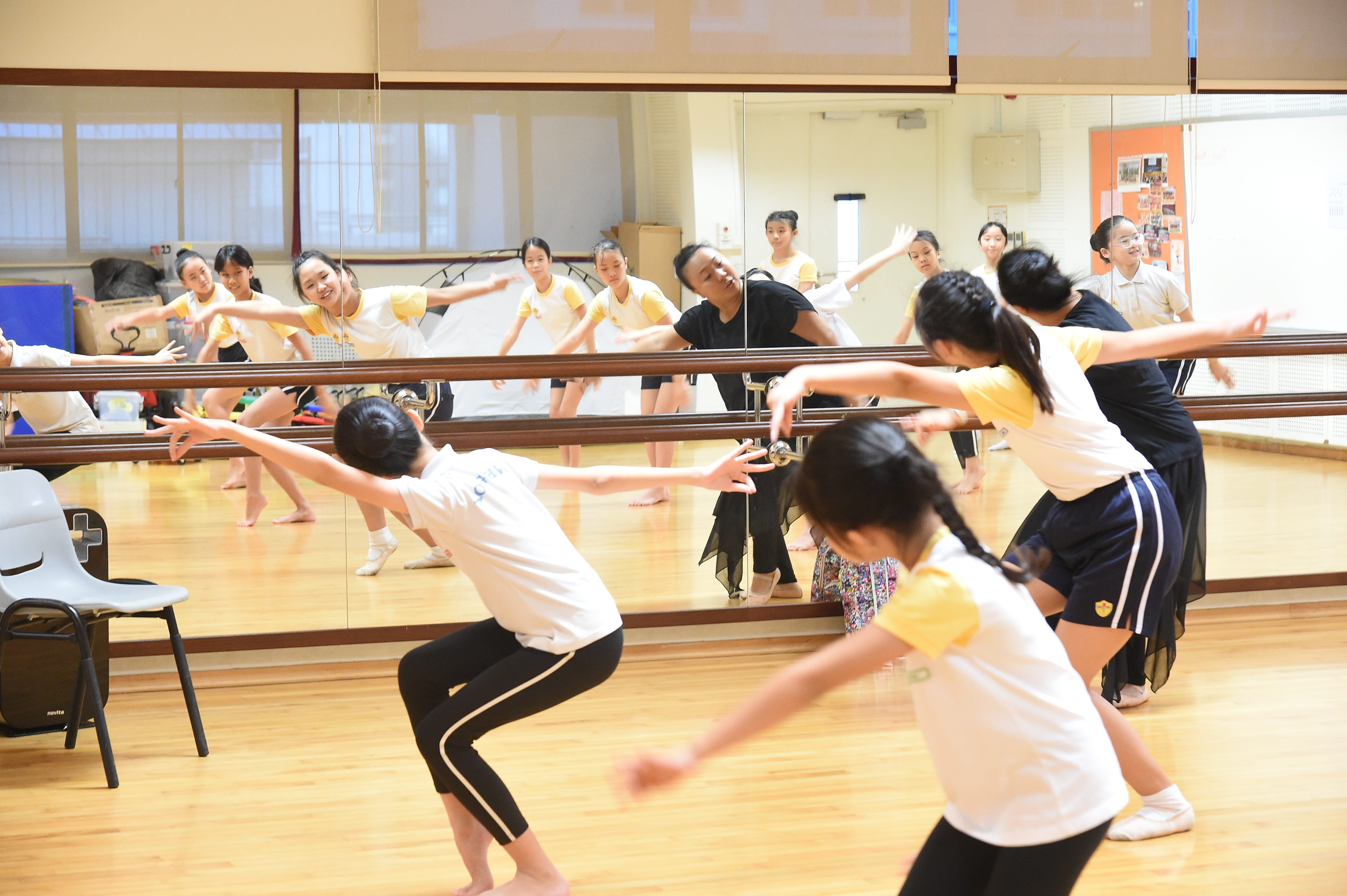 Children and an adult practice dance moves in a mirrored studio with a ballet barre along the wall.