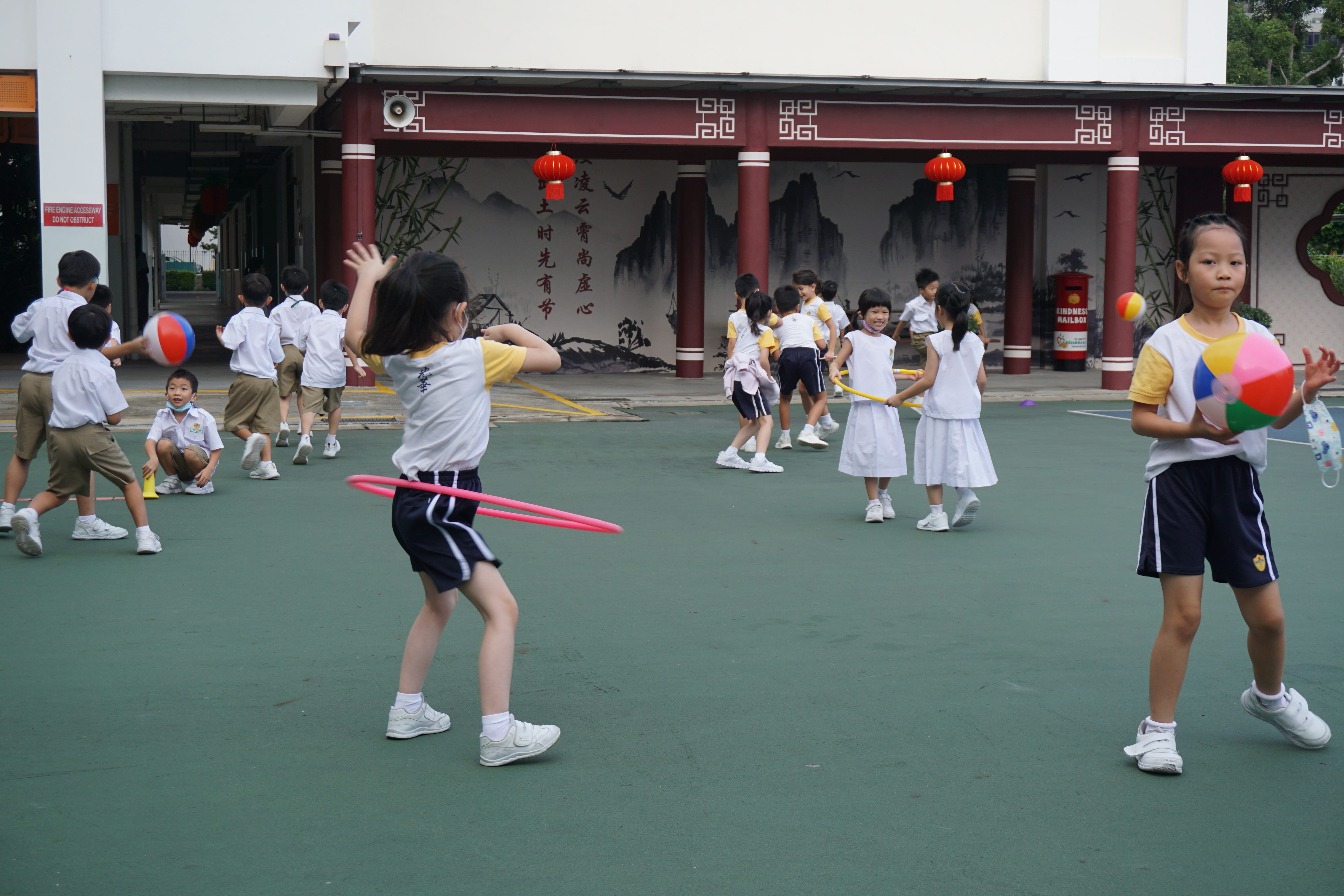 Young school children in uniforms play with green and yellow hula hoops on an outdoor court.