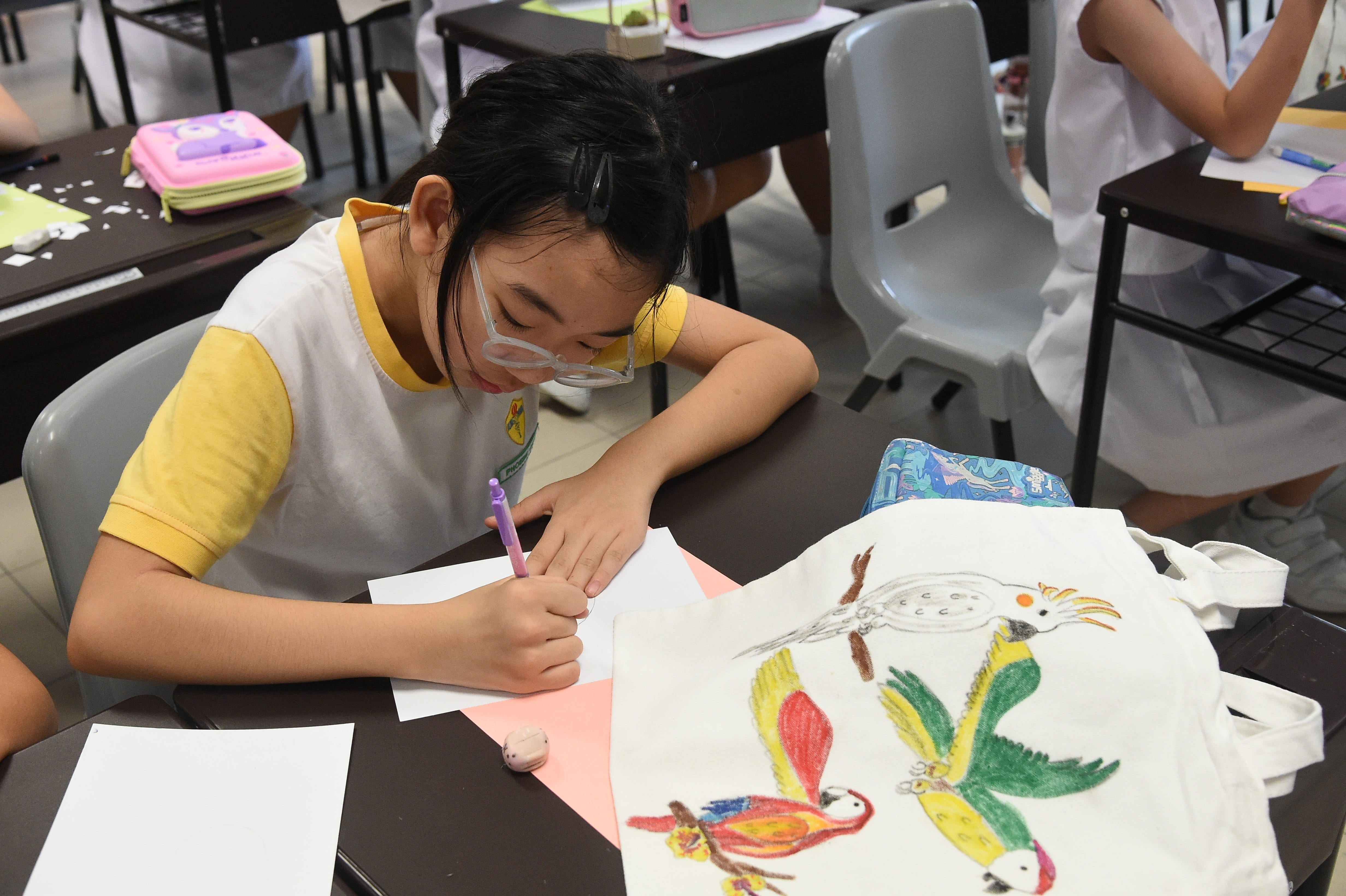 A young girl with glasses draws on paper beside a white tote bag decorated with colorful painted birds.