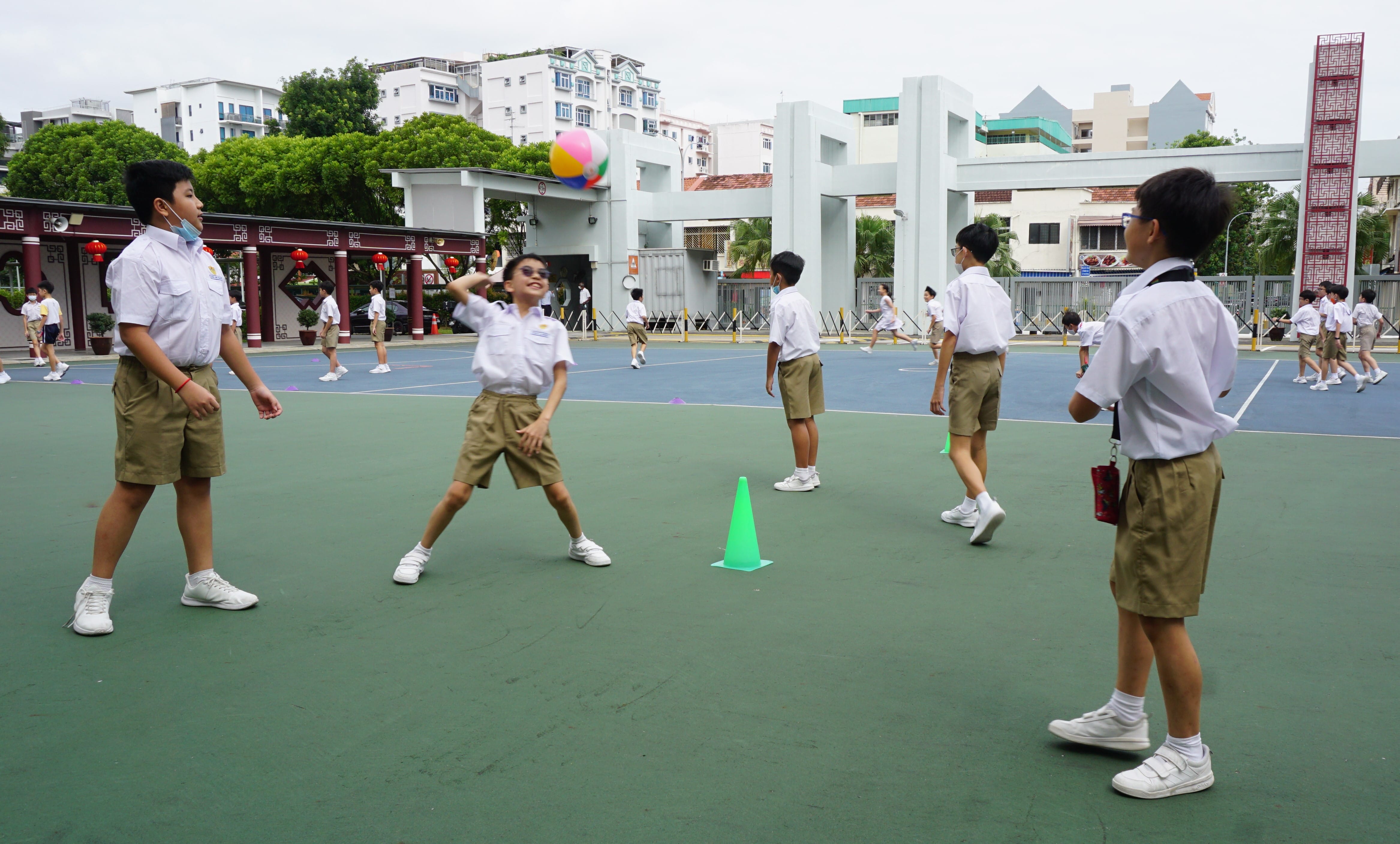 Boys in white shirts and khaki shorts play with a colorful ball on an outdoor school court with green cones.