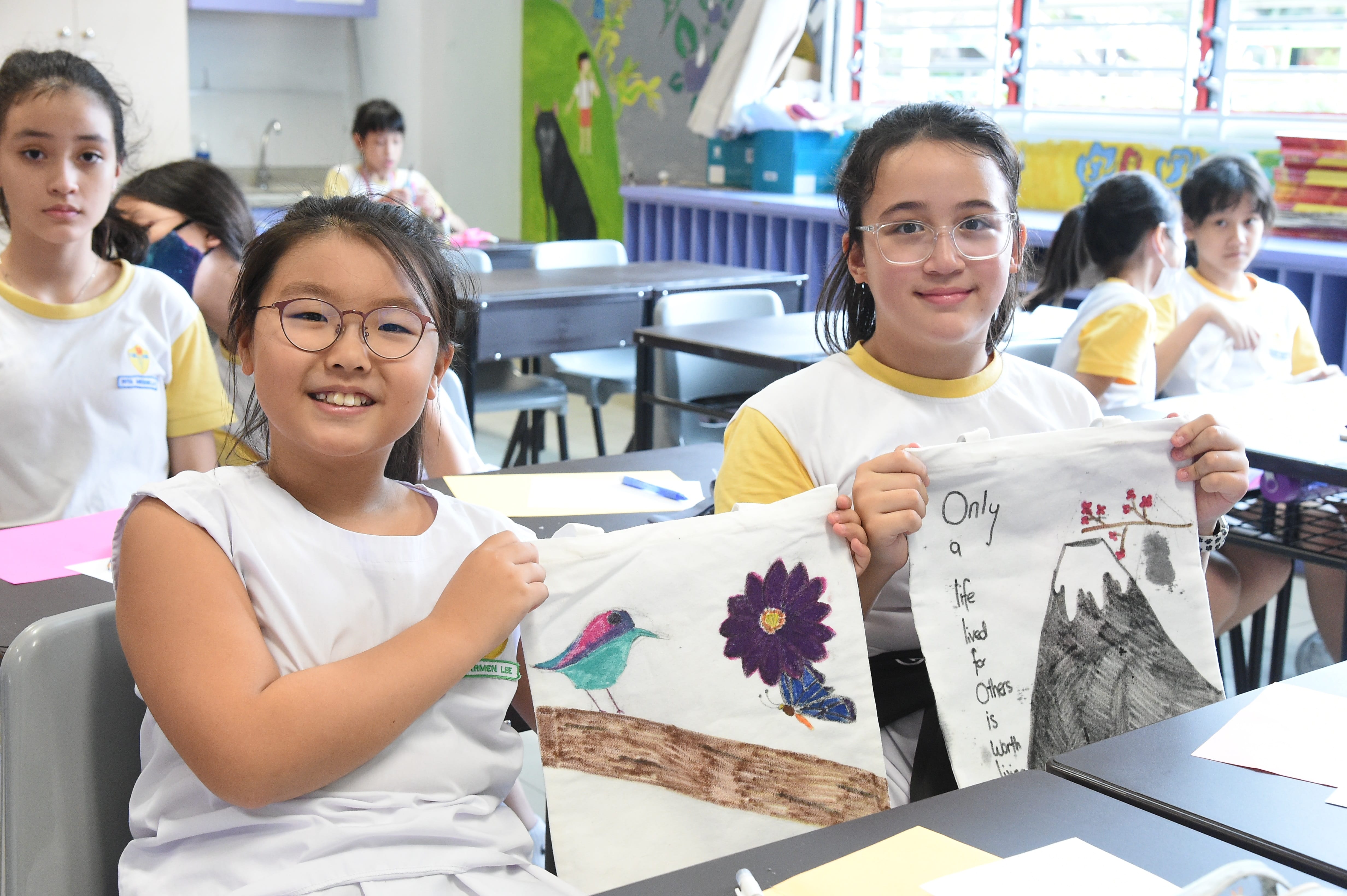 Two smiling schoolgirls hold up hand-painted fabric pieces showing a bird, flower, and motivational quote in a classroom.