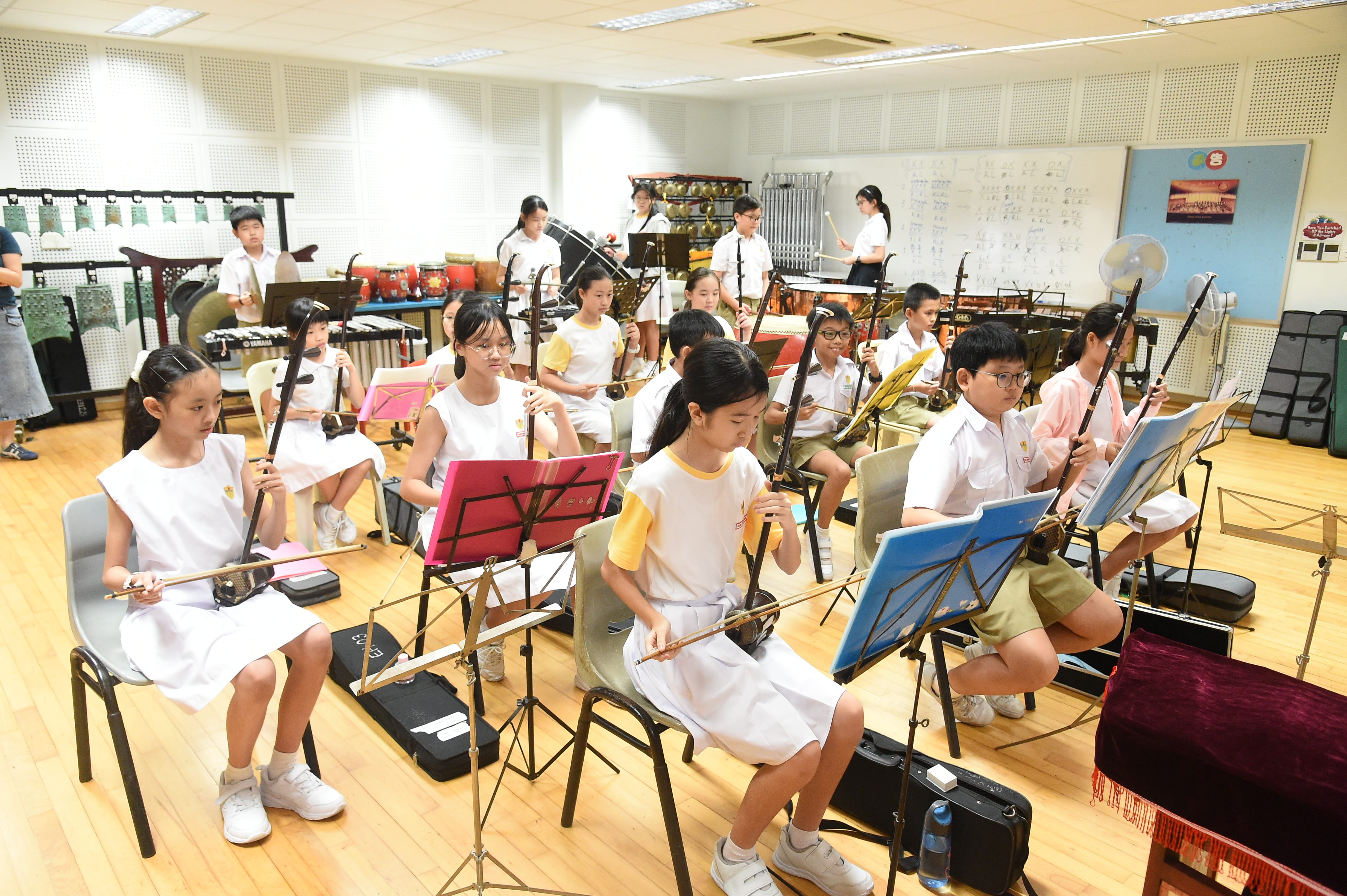 Children in school uniforms sit in a music room playing erhu instruments with sheet music on stands.