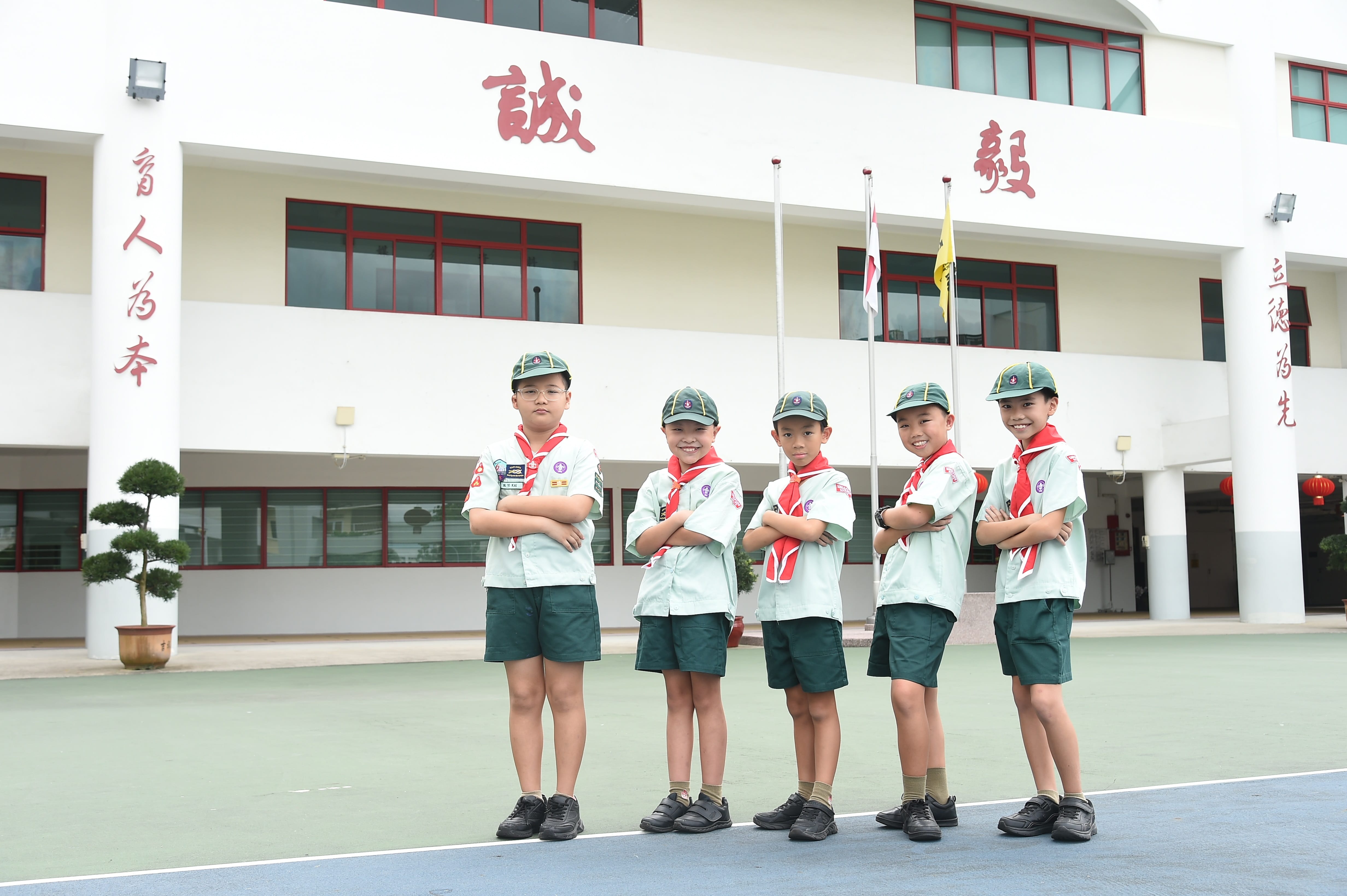 Five children in scout uniforms with red scarves and green caps stand arms crossed in front of a white school building with Chinese characters.