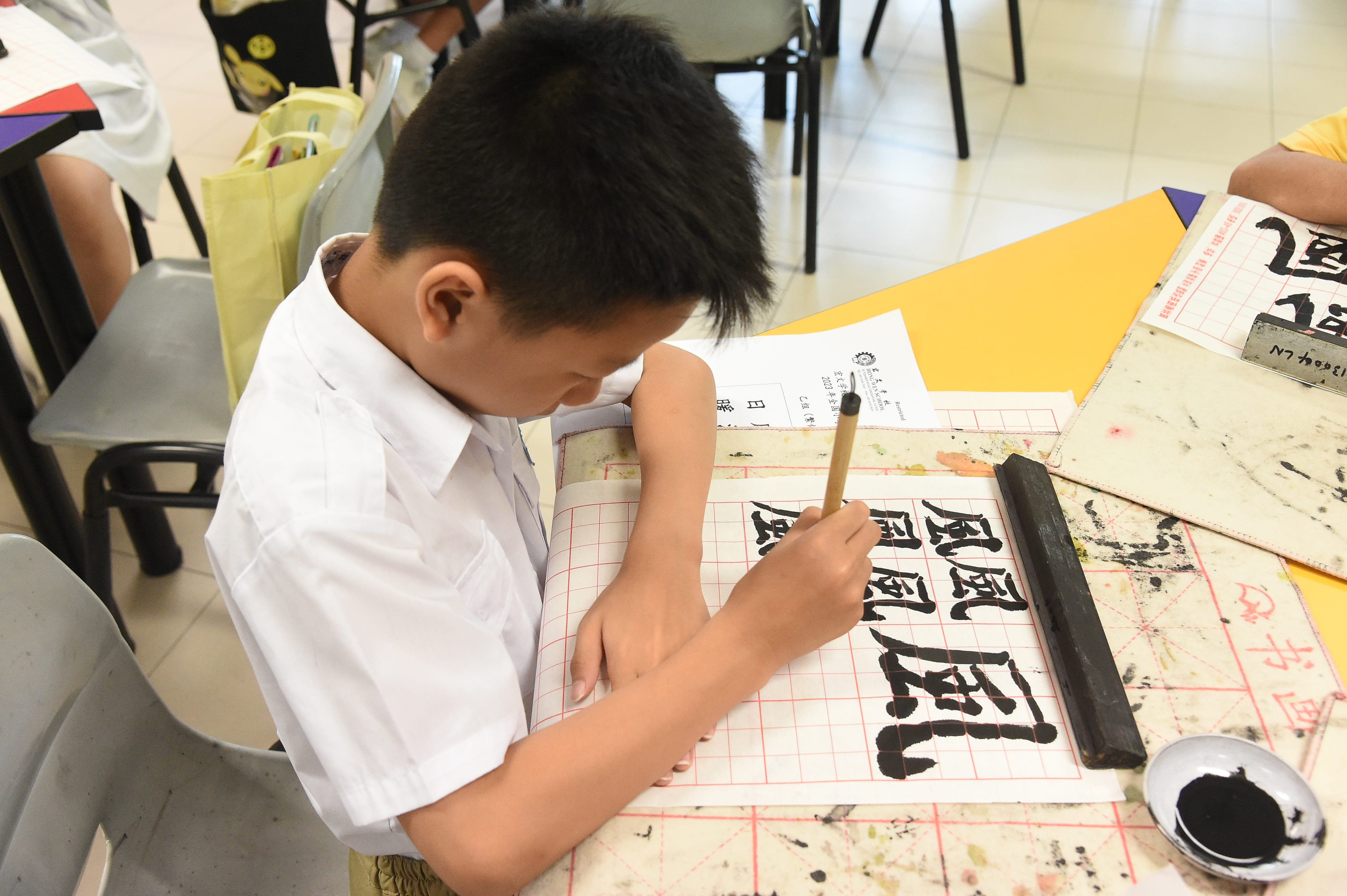 A young boy in a white shirt practices Chinese calligraphy with a brush and black ink on grid paper.