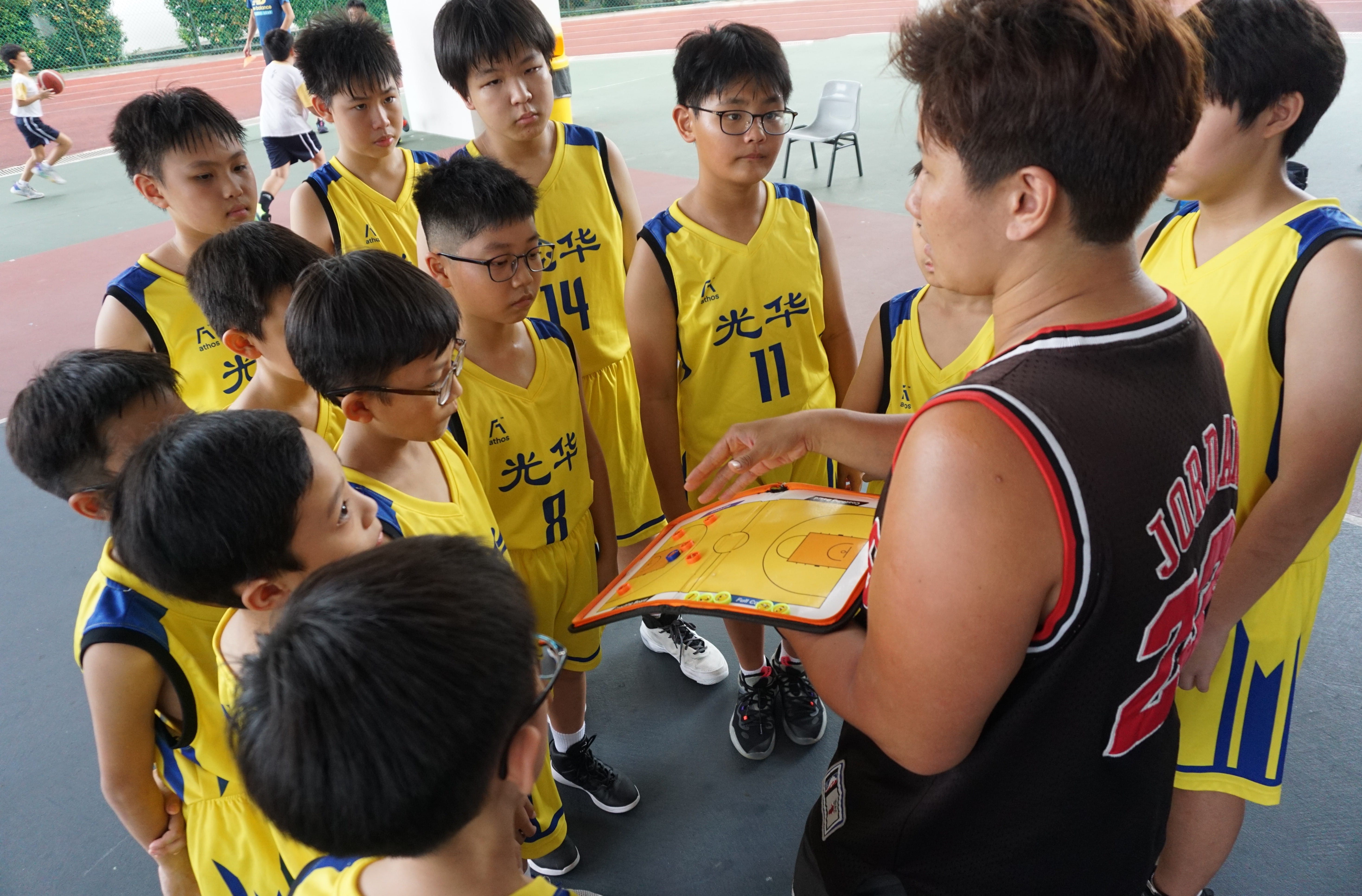 A coach shows a tactical board to young basketball players in yellow uniforms on an outdoor court.