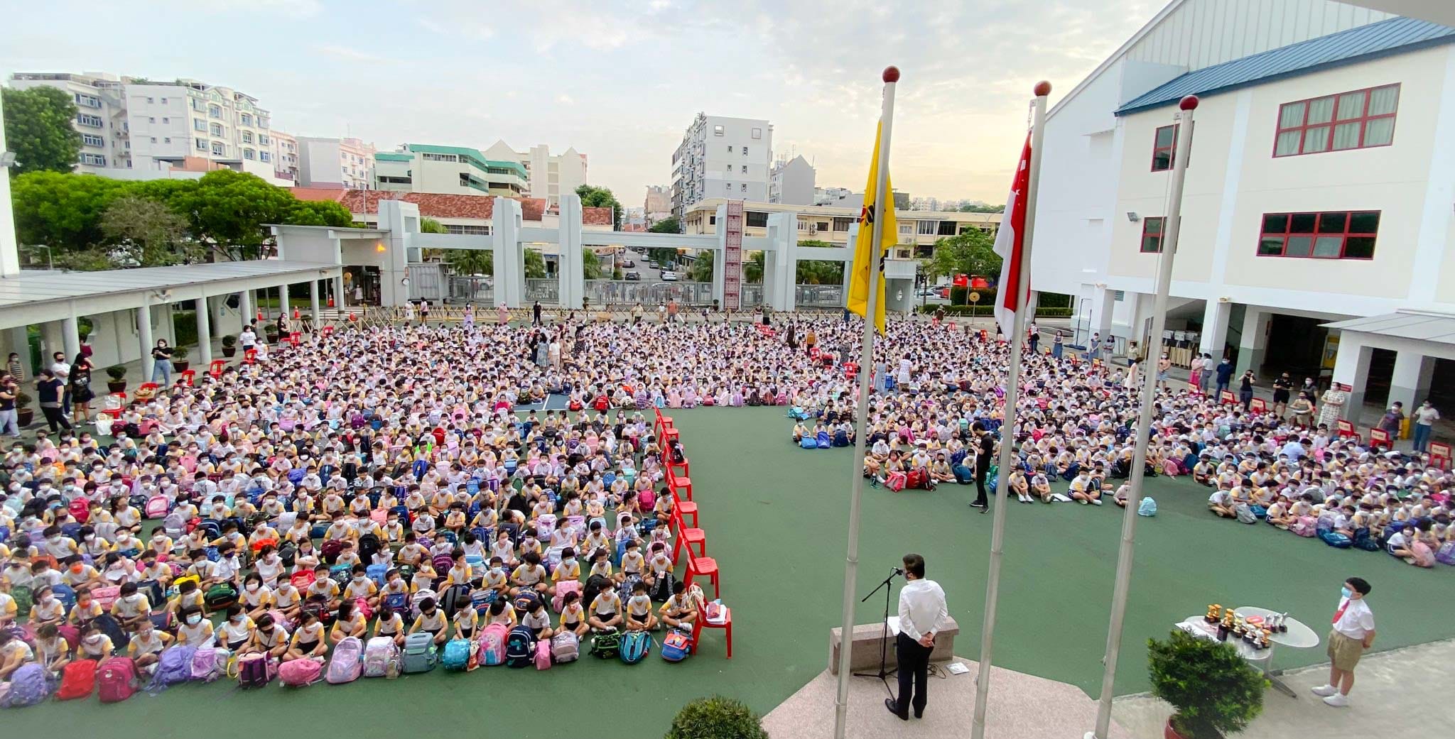 Hundreds of masked students sit on a school courtyard during an outdoor assembly with a speaker at a podium.