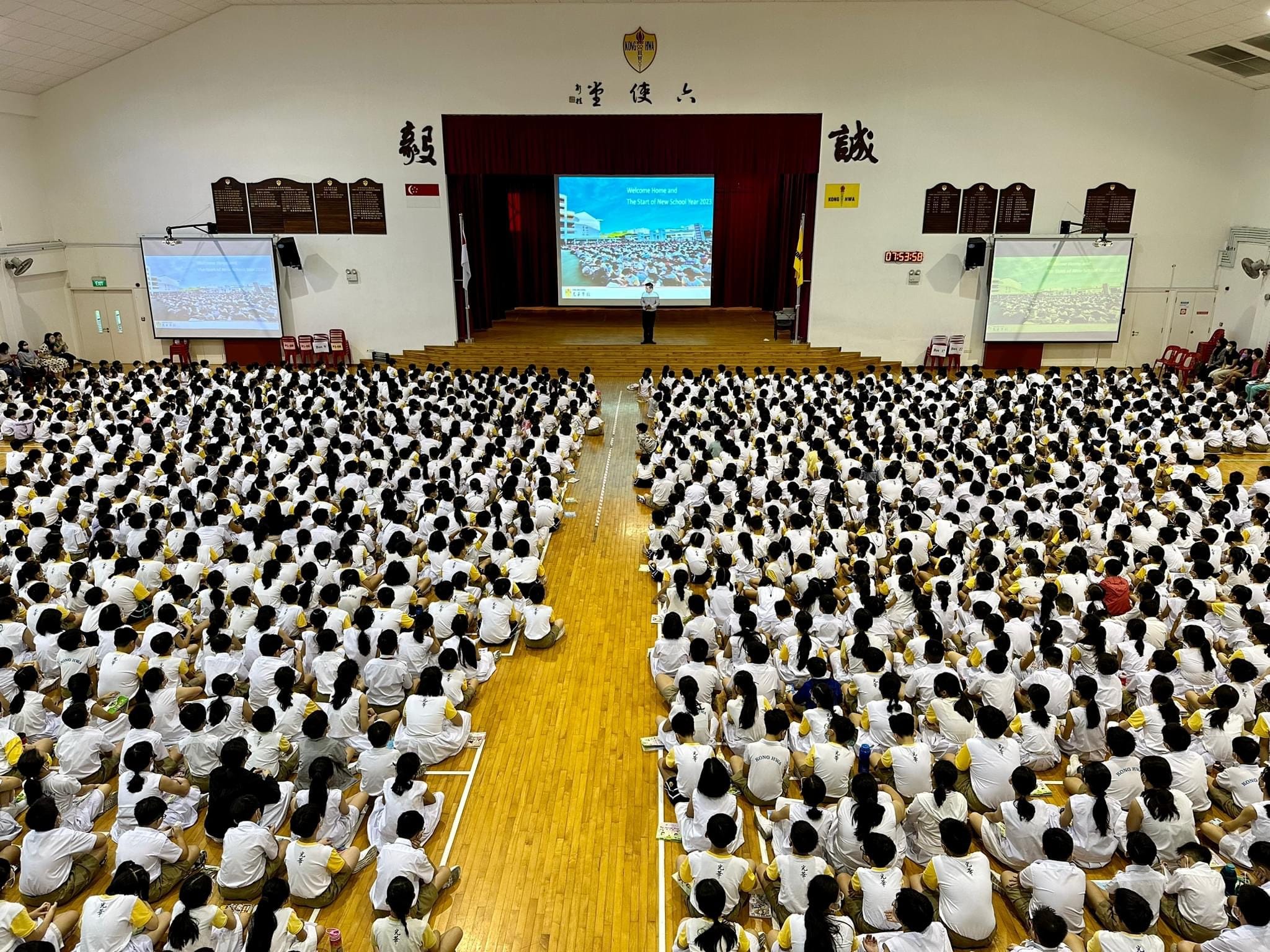 Hundreds of students in white uniforms sit on a school hall floor facing a stage with a projected welcome screen.