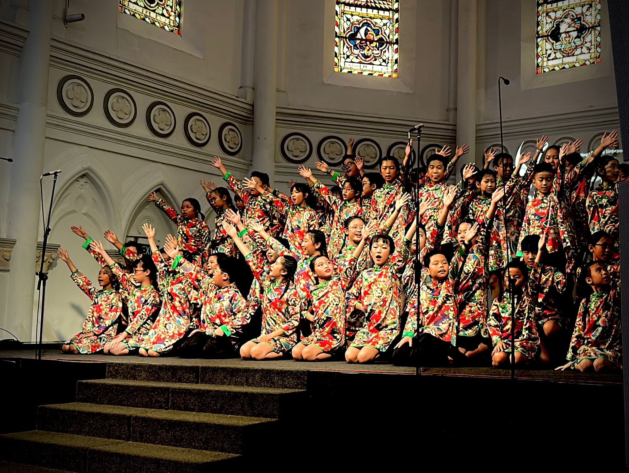 Children in colorful floral robes perform a choir concert on church steps with stained glass windows behind them.