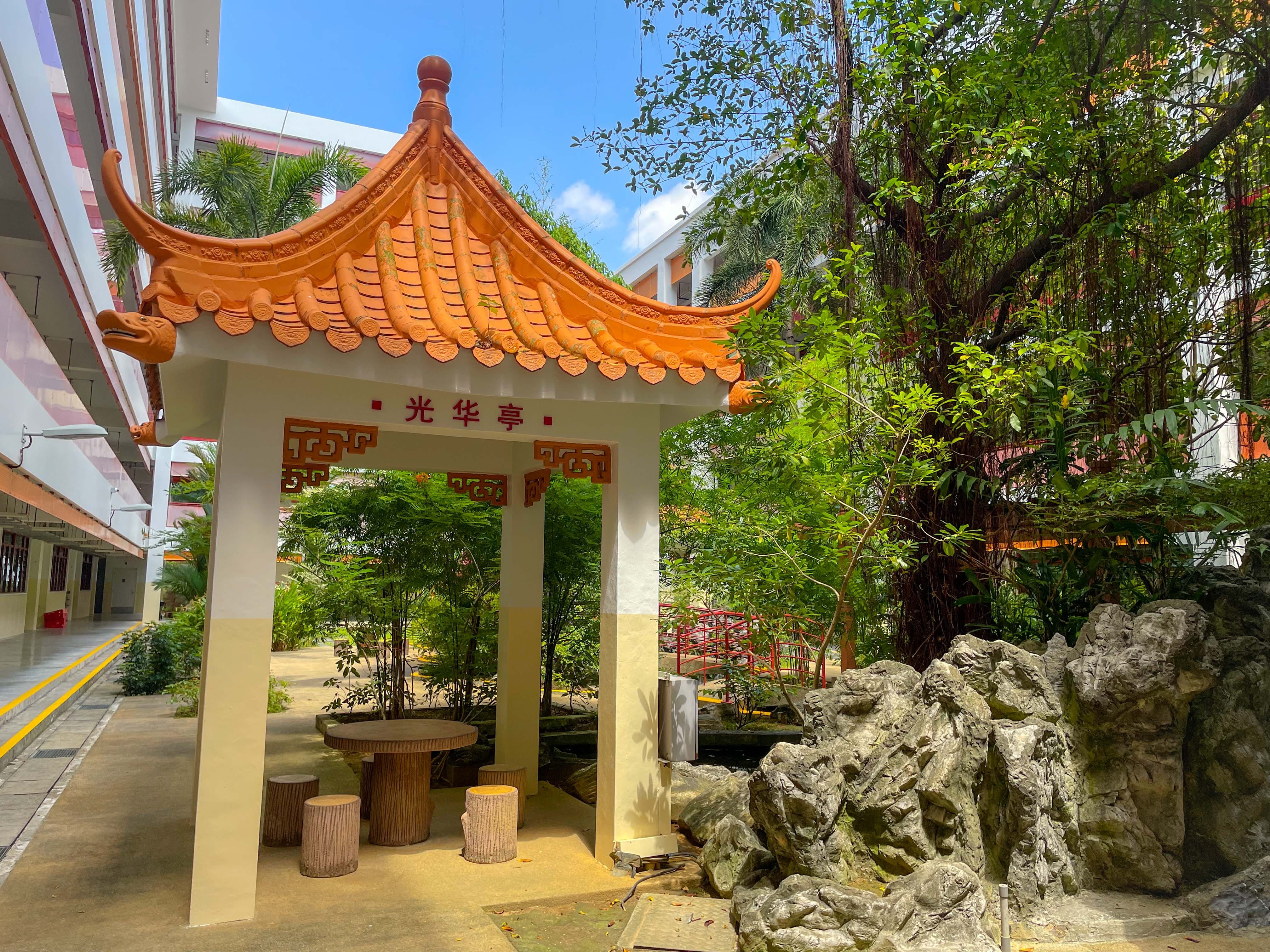 Orange-roofed Chinese pavilion with red characters, stone seats, rock formation, and green trees beside a building.