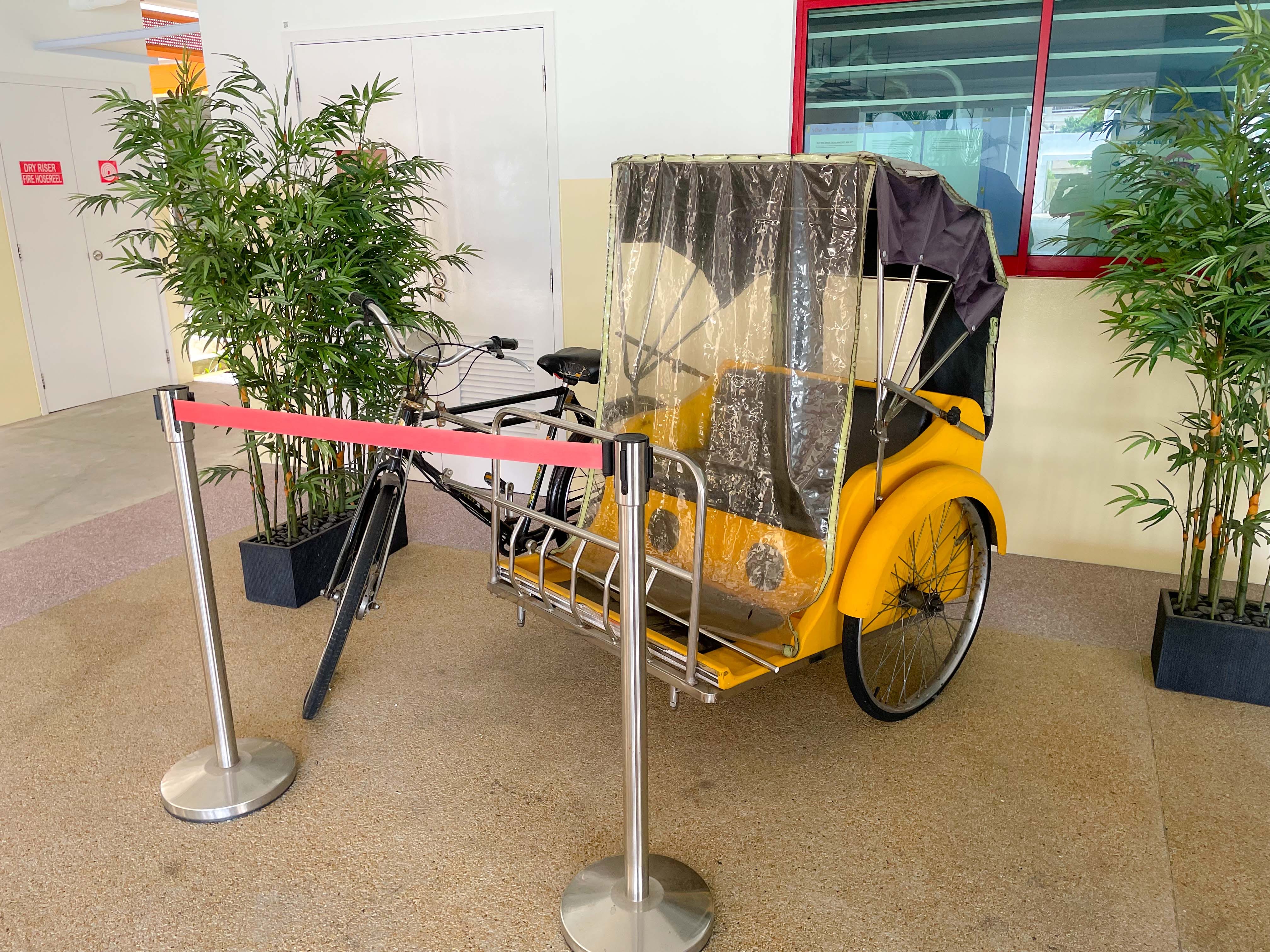 A yellow pedicab rickshaw with a clear plastic canopy on display indoors, surrounded by bamboo plants and red barrier ropes.