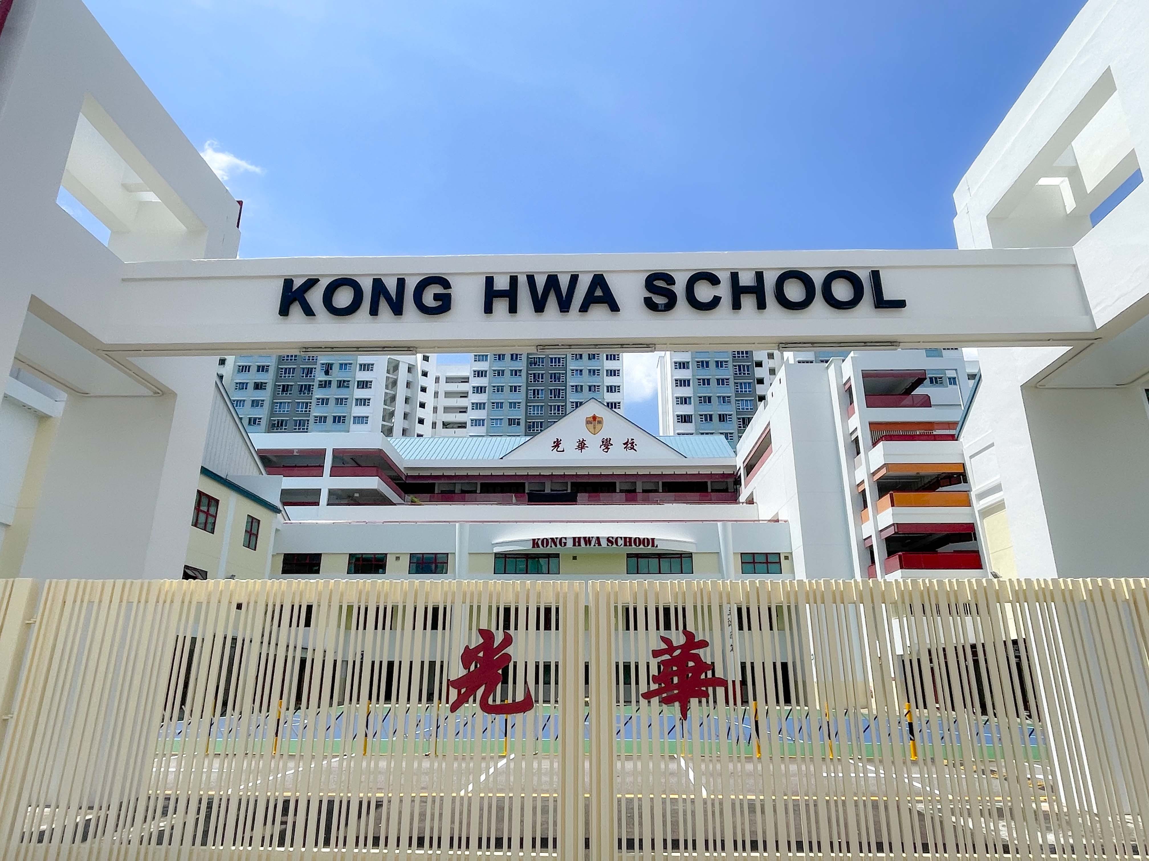White entrance gate of Kong Hwa School with Chinese characters on the fence and school buildings behind under blue sky.