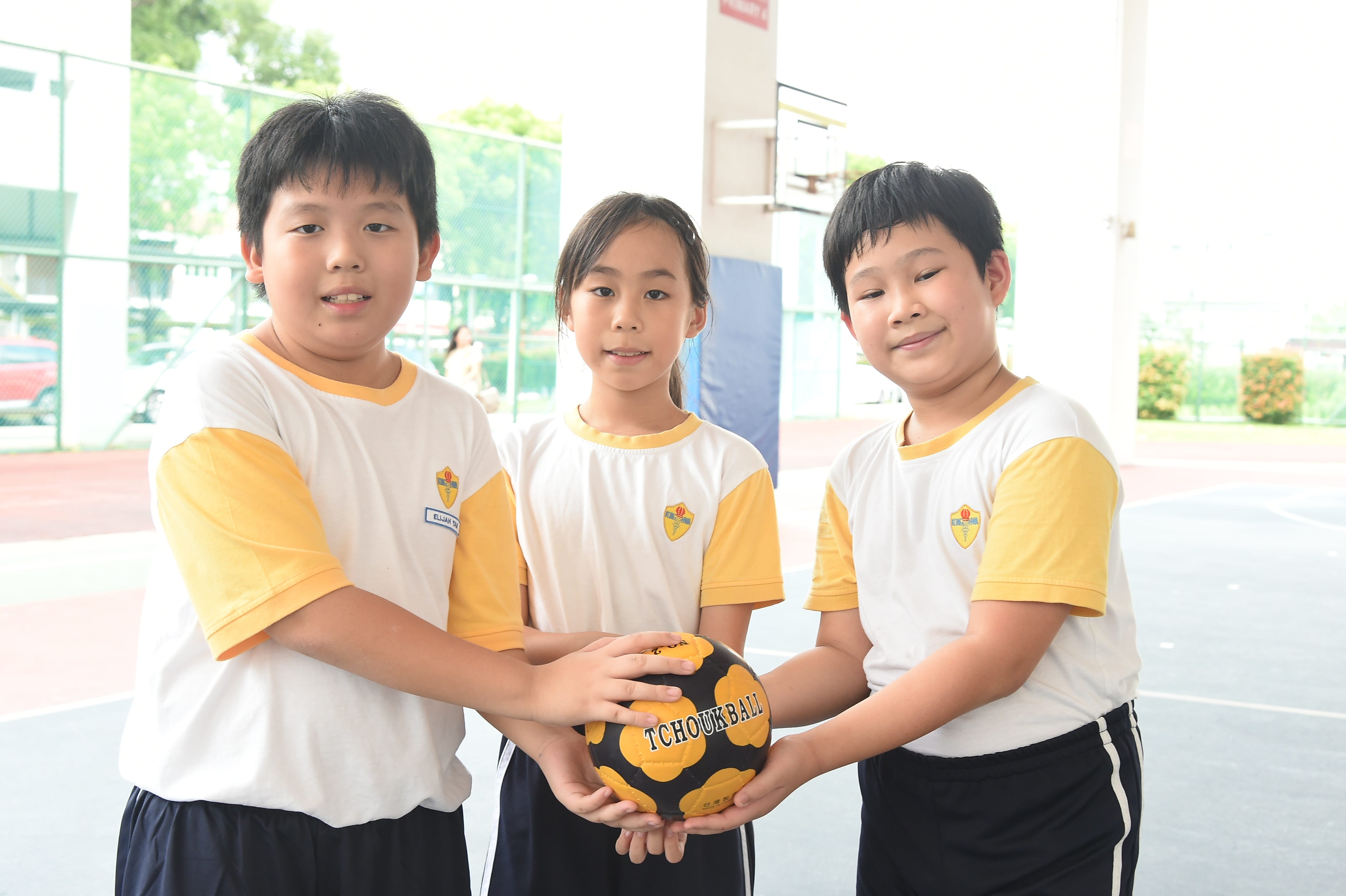 Three children in matching school uniforms hold a black and yellow tchoukball together on an outdoor court.